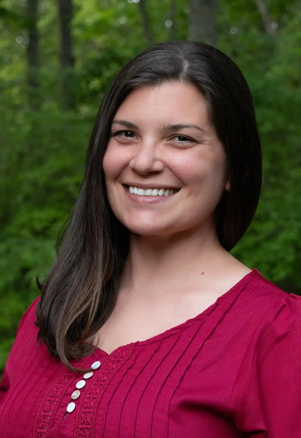 A woman with long dark hair smiling outdoors in front of a green forest background, wearing a red top.