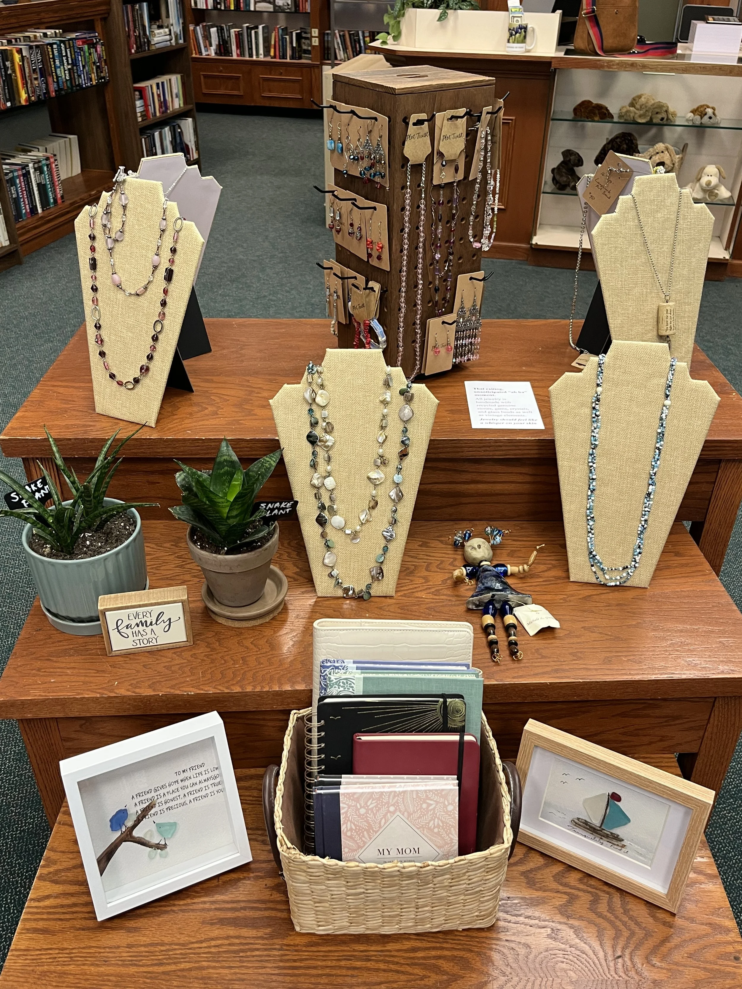 A display of jewelry including necklaces, bracelets, and earrings, arranged on neck busts and a wooden stand in a store. There are also framed pictures, a small potted plant, and a basket with notebooks on the table.
