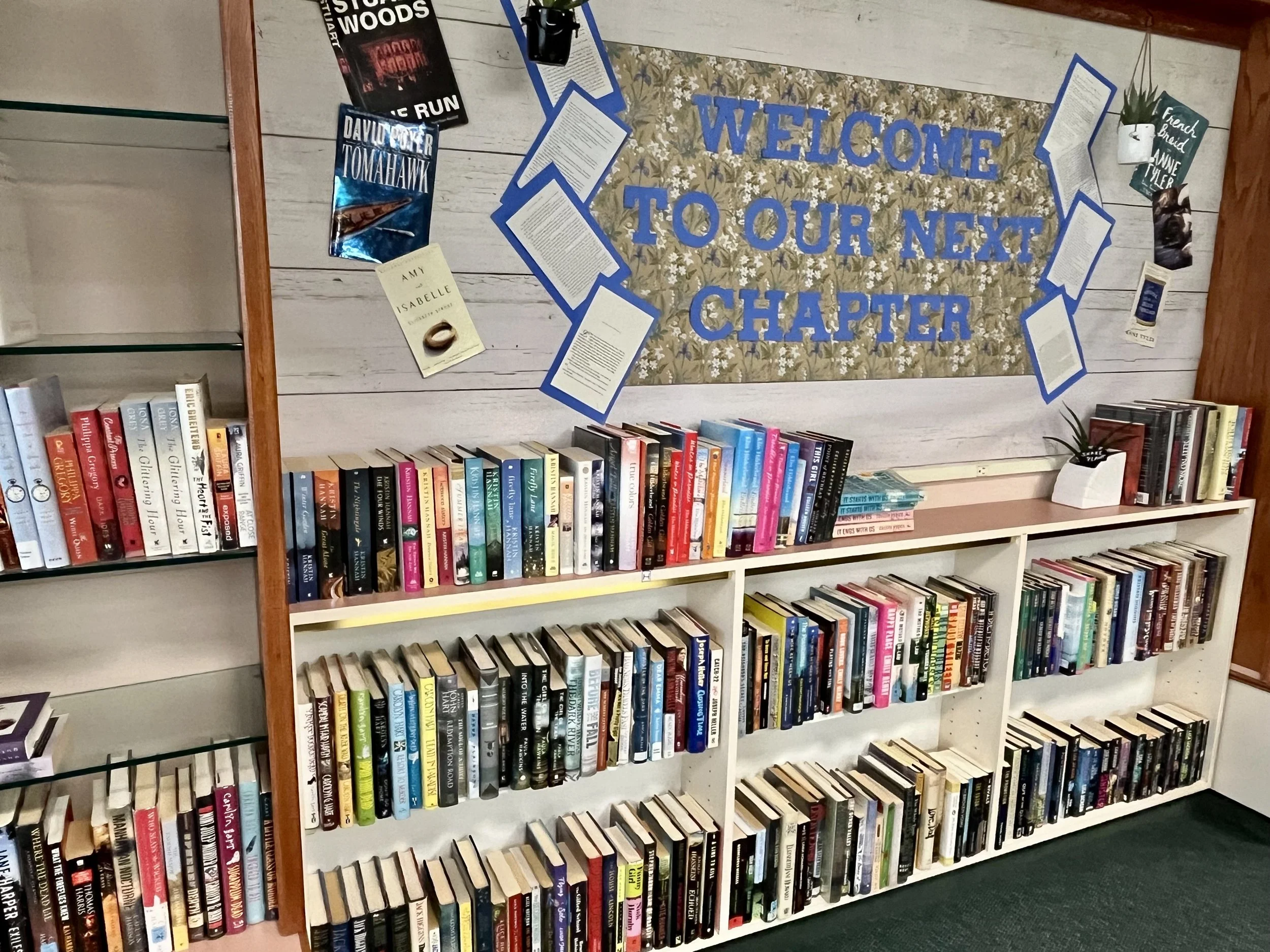 Bookshelves filled with books and a decorative welcome sign on a wall with blue letter cutouts that says, 'Welcome to Our Next Chapter.'