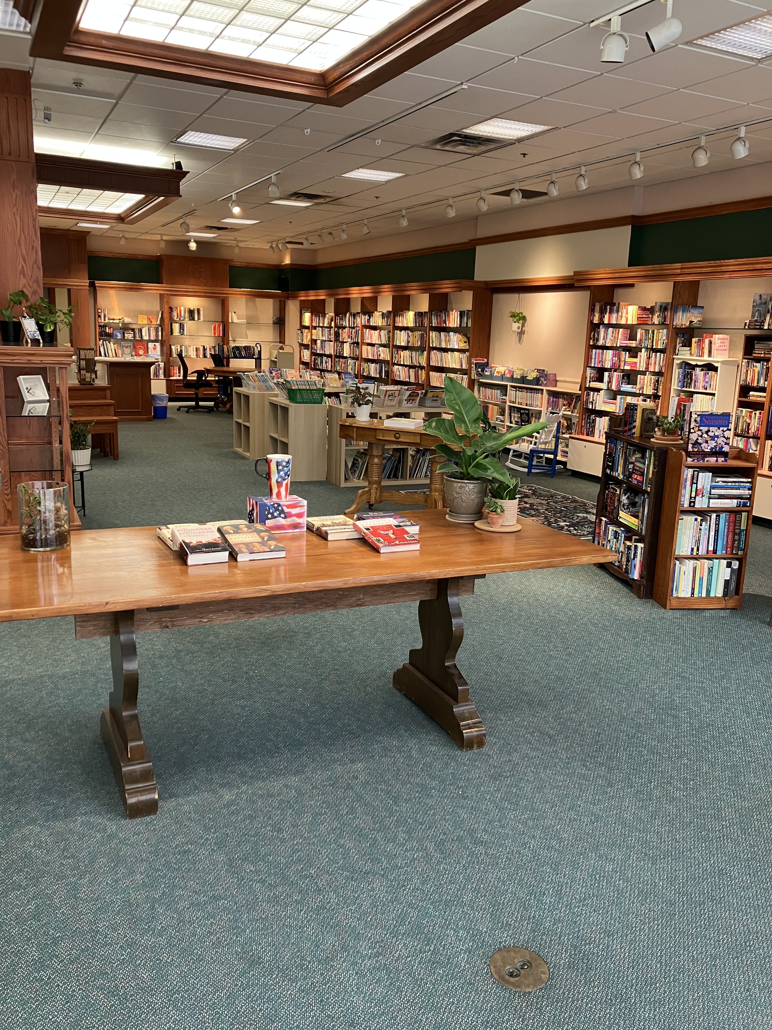 A cozy library with wooden bookshelves filled with books, a wooden table with books and plants, and green carpeted floors.