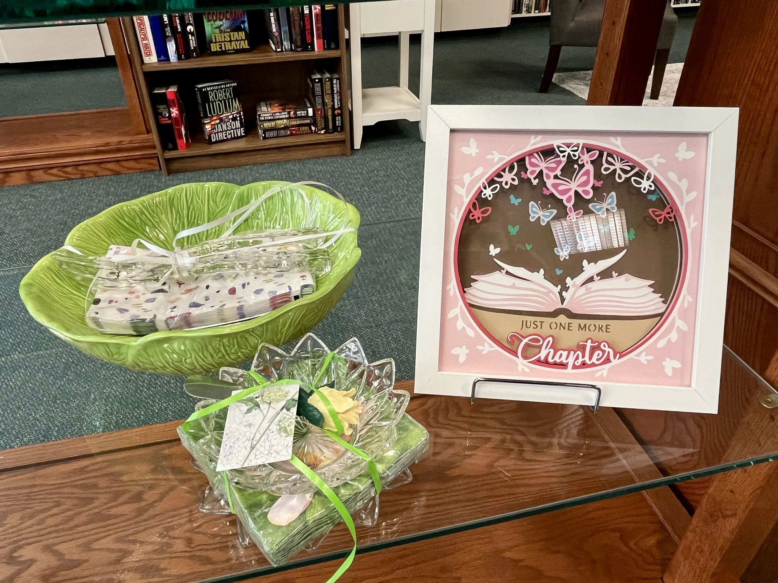 Bookshelf with various books, a framed decorative wall art with pink, blue, and white butterflies around an open book illustration, and a green basket or container with decorative items on a wooden surface.