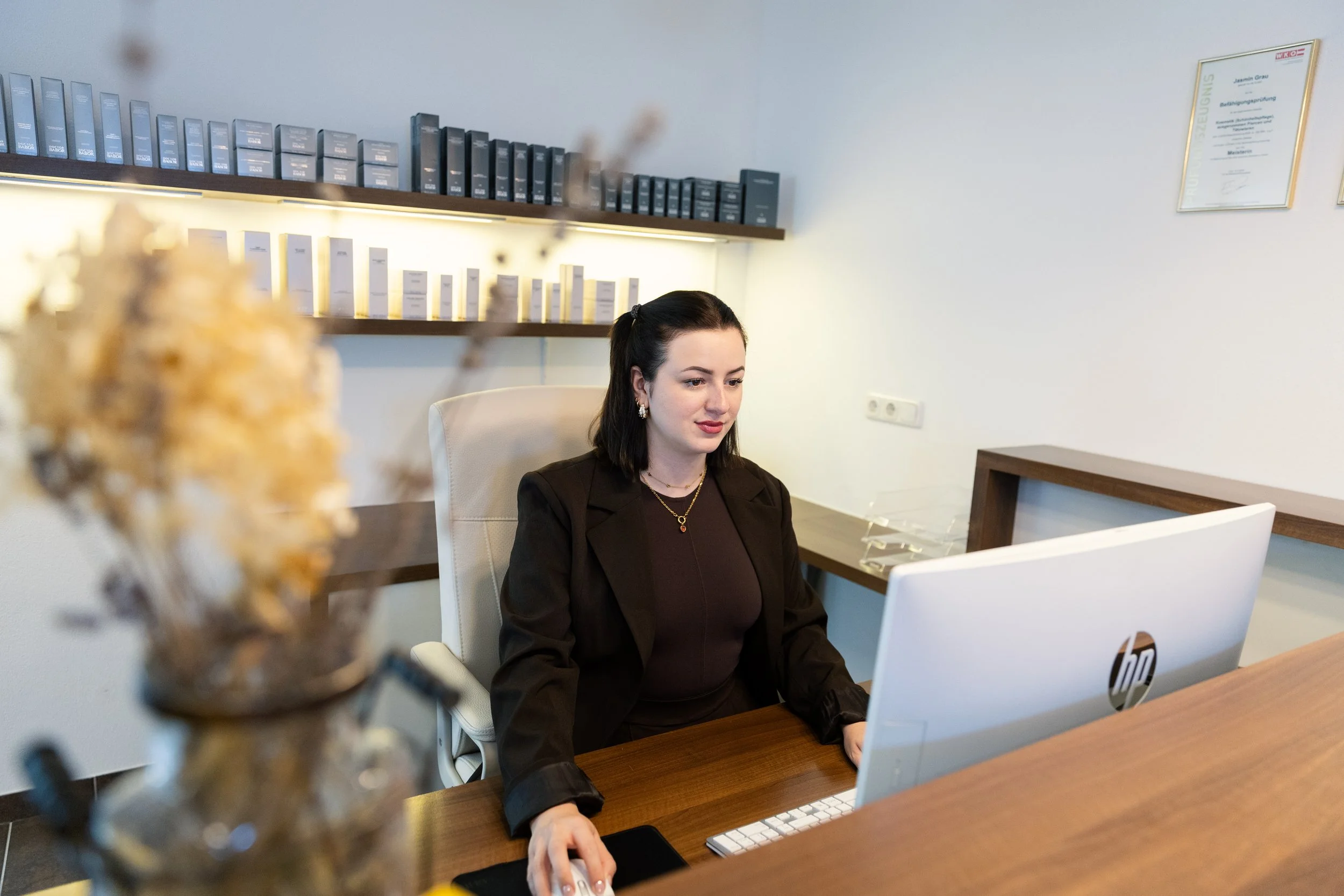 Eine Frau sitzt an einem Schreibtisch vor einem HP-Desktopcomputer, gearbeitet in einem Büro mit weißen Wänden, Regalen mit Aktenordnern und einem DIN-A4-Zeugnis an der Wand.