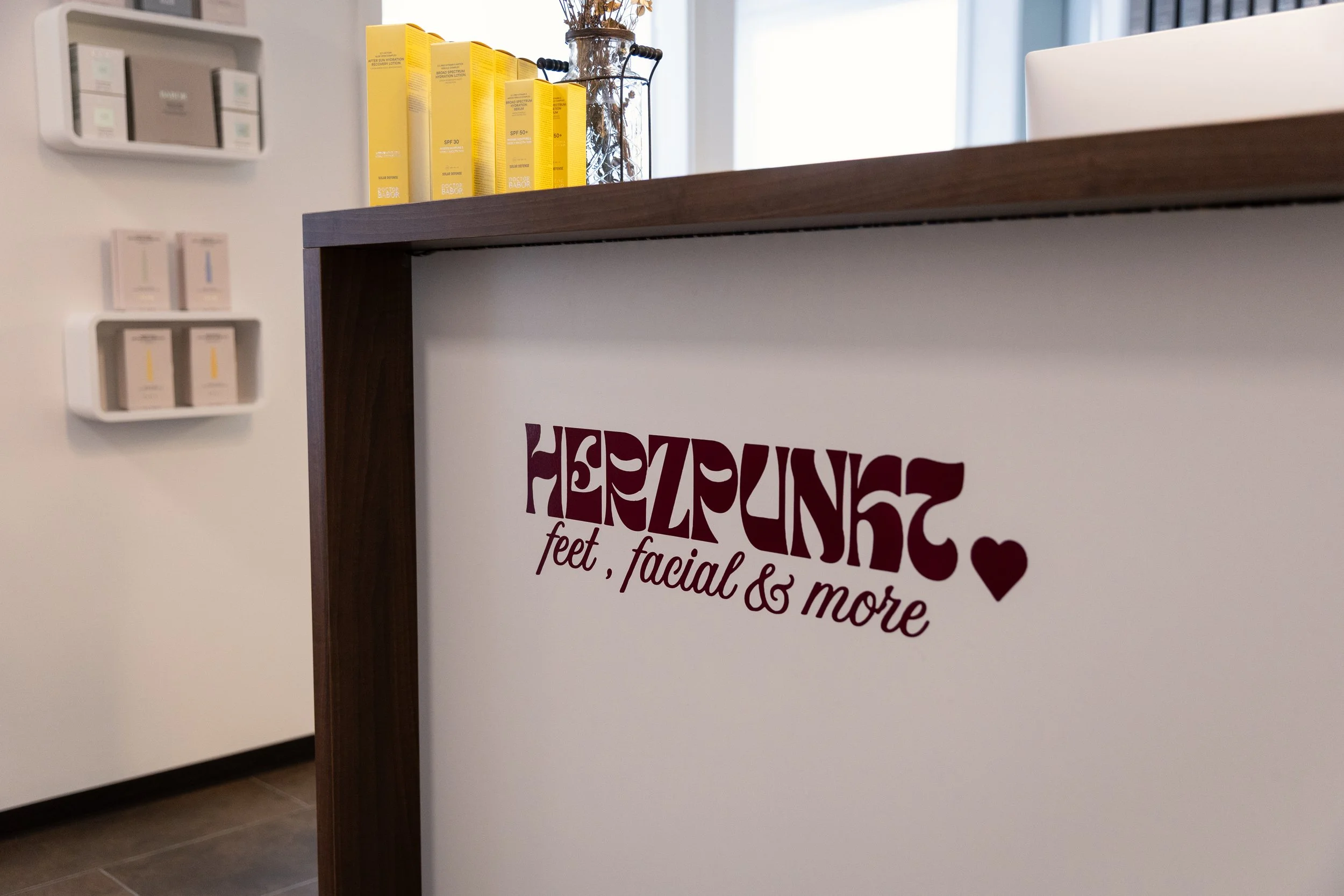 Ein Bild eines Innenraums, das eine Theke mit einem Schild zeigt, auf dem steht »feet, facial & more«. Im Hintergrund sind Regale mit verschiedenen Hautpflegeprodukten und eine Vase mit Zweigen.