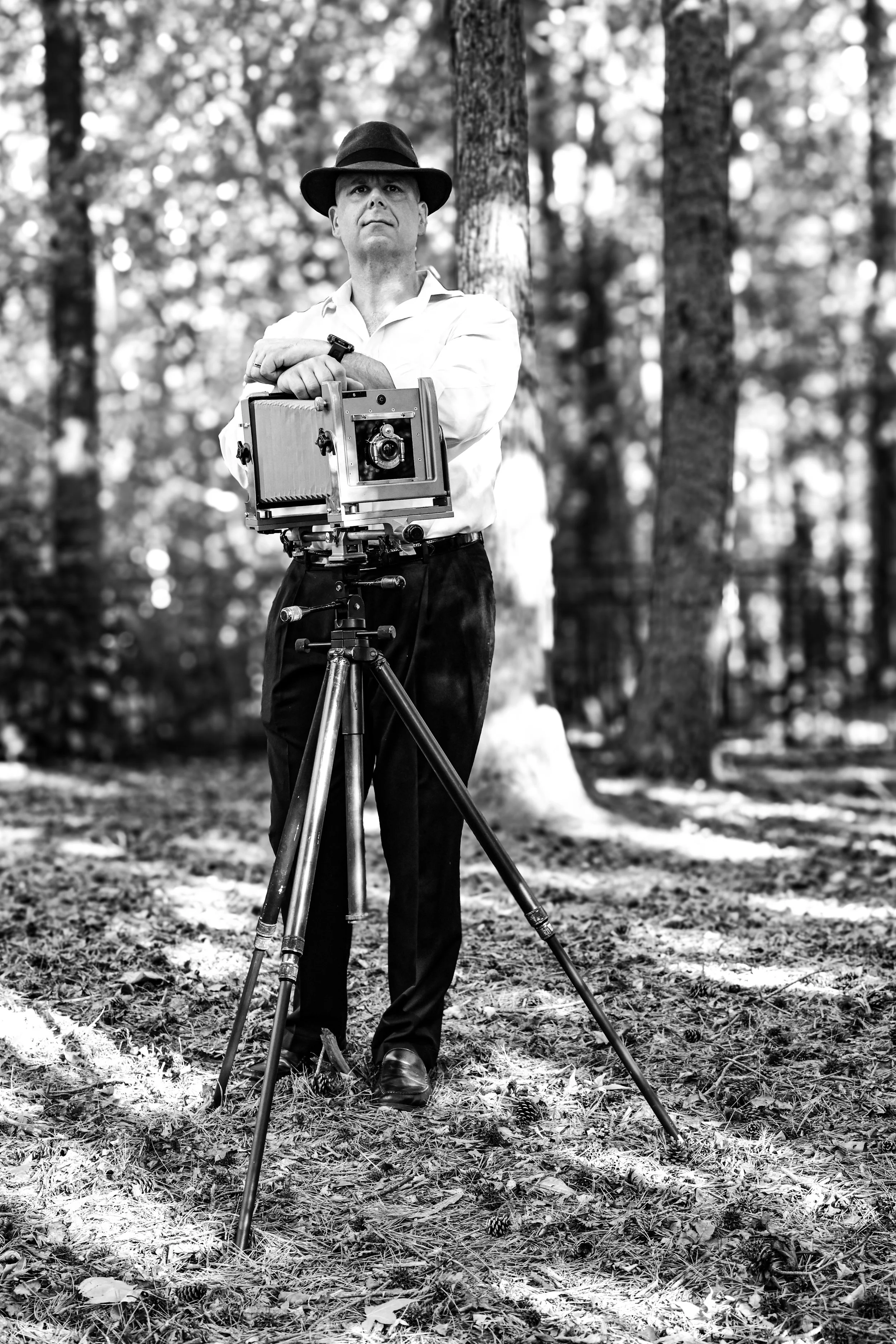 Black and white photo of a man standing outdoors in a forest, adjusting a large camera mounted on a tripod, wearing a fedora hat and a white shirt.