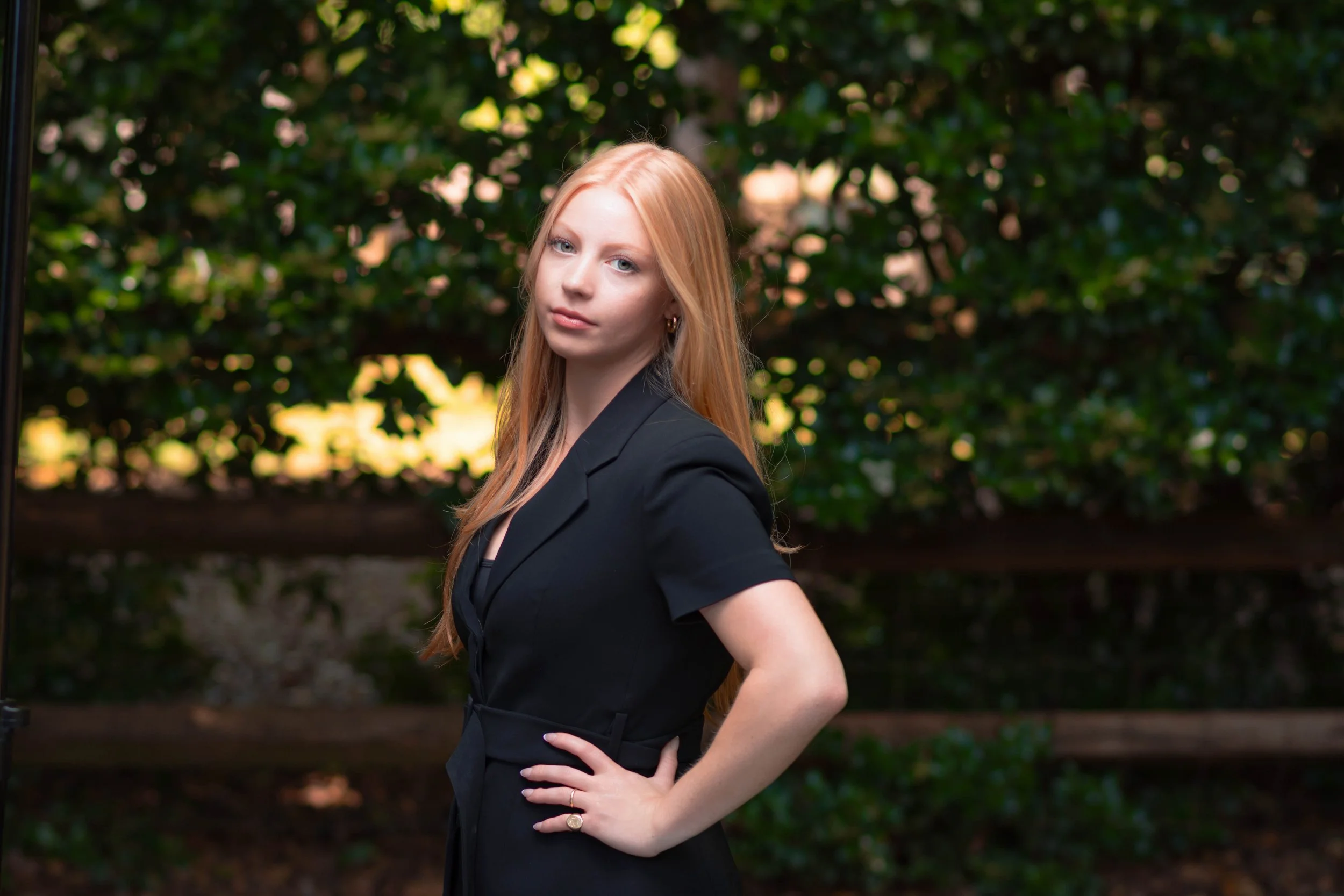 A young woman with long red hair wearing a black business suit, standing outdoors in front of a green leafy background, with her hand on her hip and looking into the camera with a confident expression.