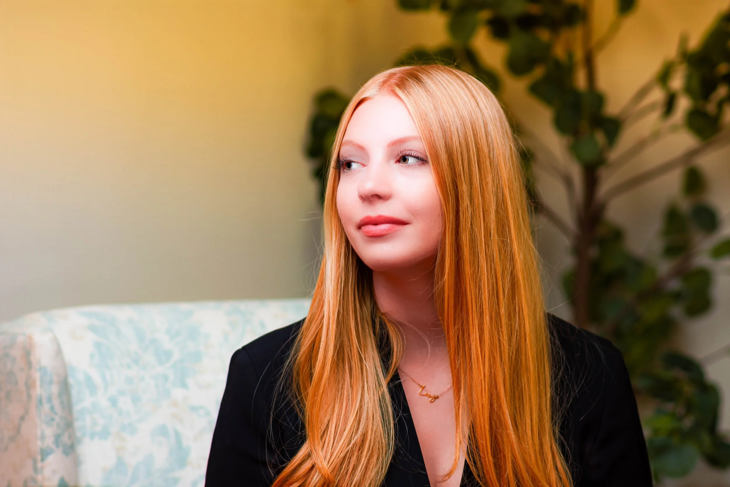 A young woman with long red hair looking to her right, wearing a black top and a gold necklace, sitting in a room with a plant and a light-colored wall in the background.