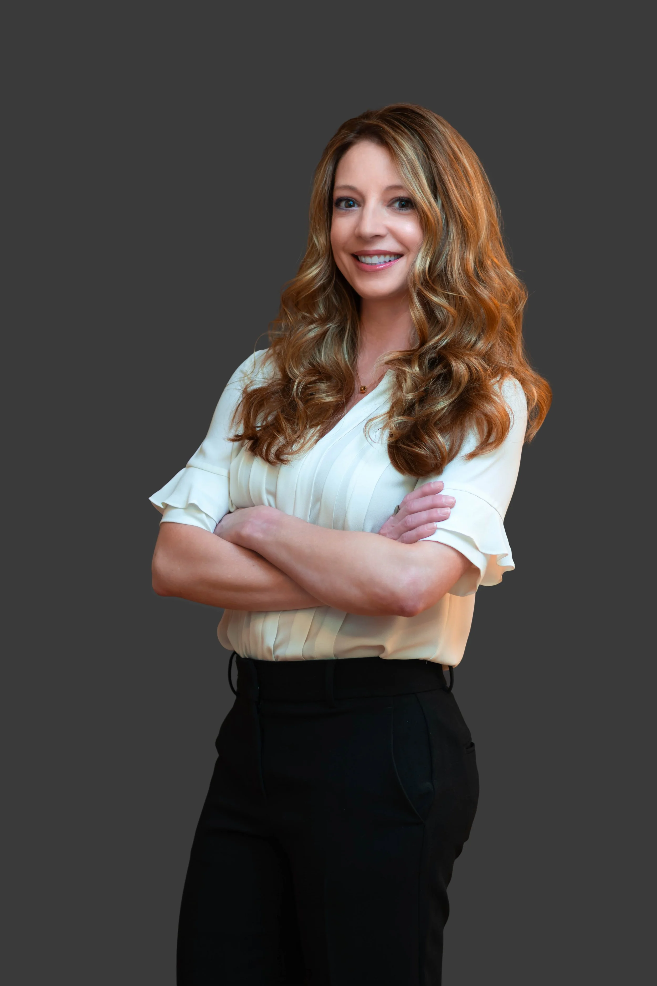 A smiling woman with long, wavy red hair wearing a cream-colored blouse and black pants, standing with her arms crossed against a plain dark background.