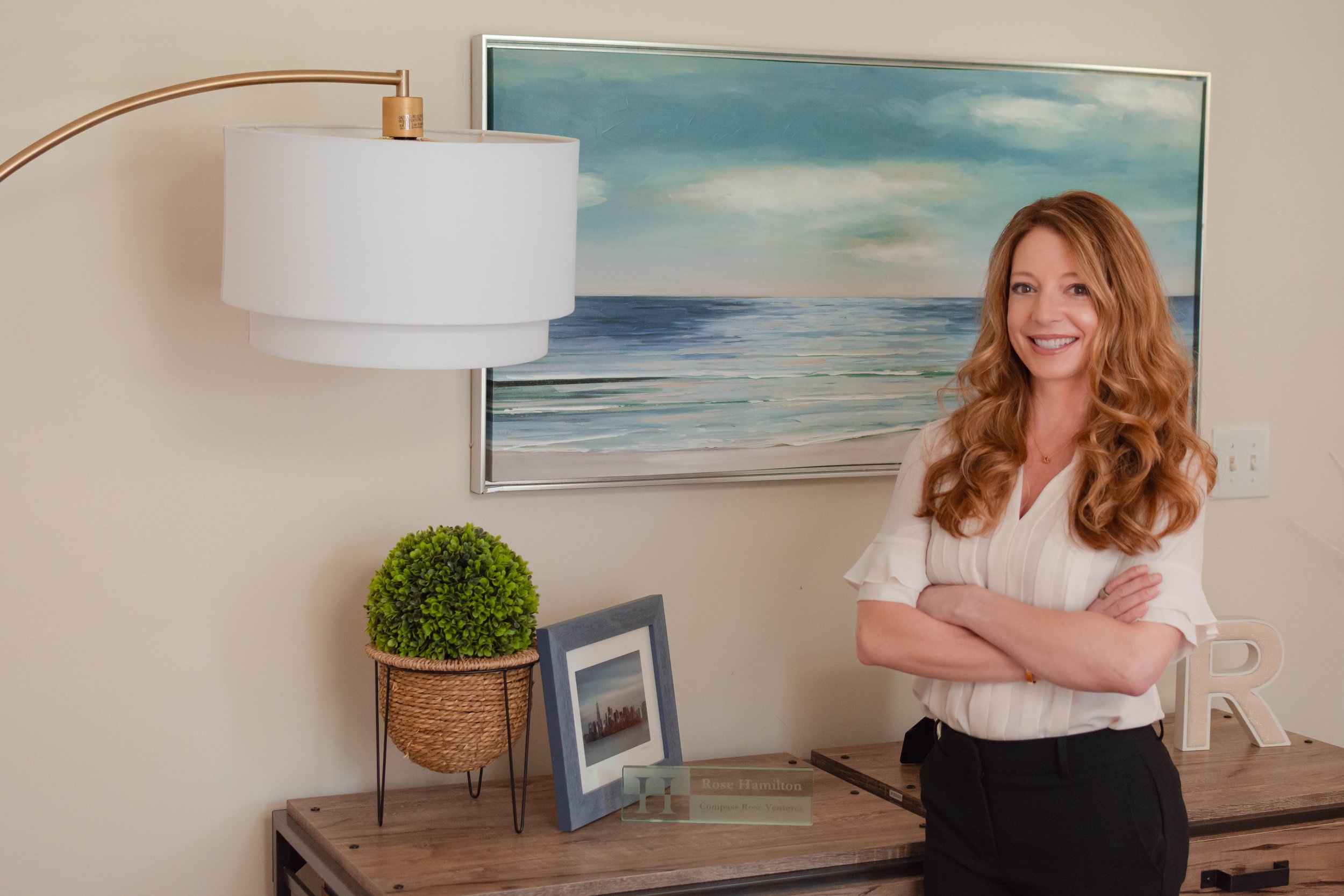 A woman with curly red hair smiling with arms crossed in a room with wall art, a lamp, a potted plant, and framed photos on a wooden table.
