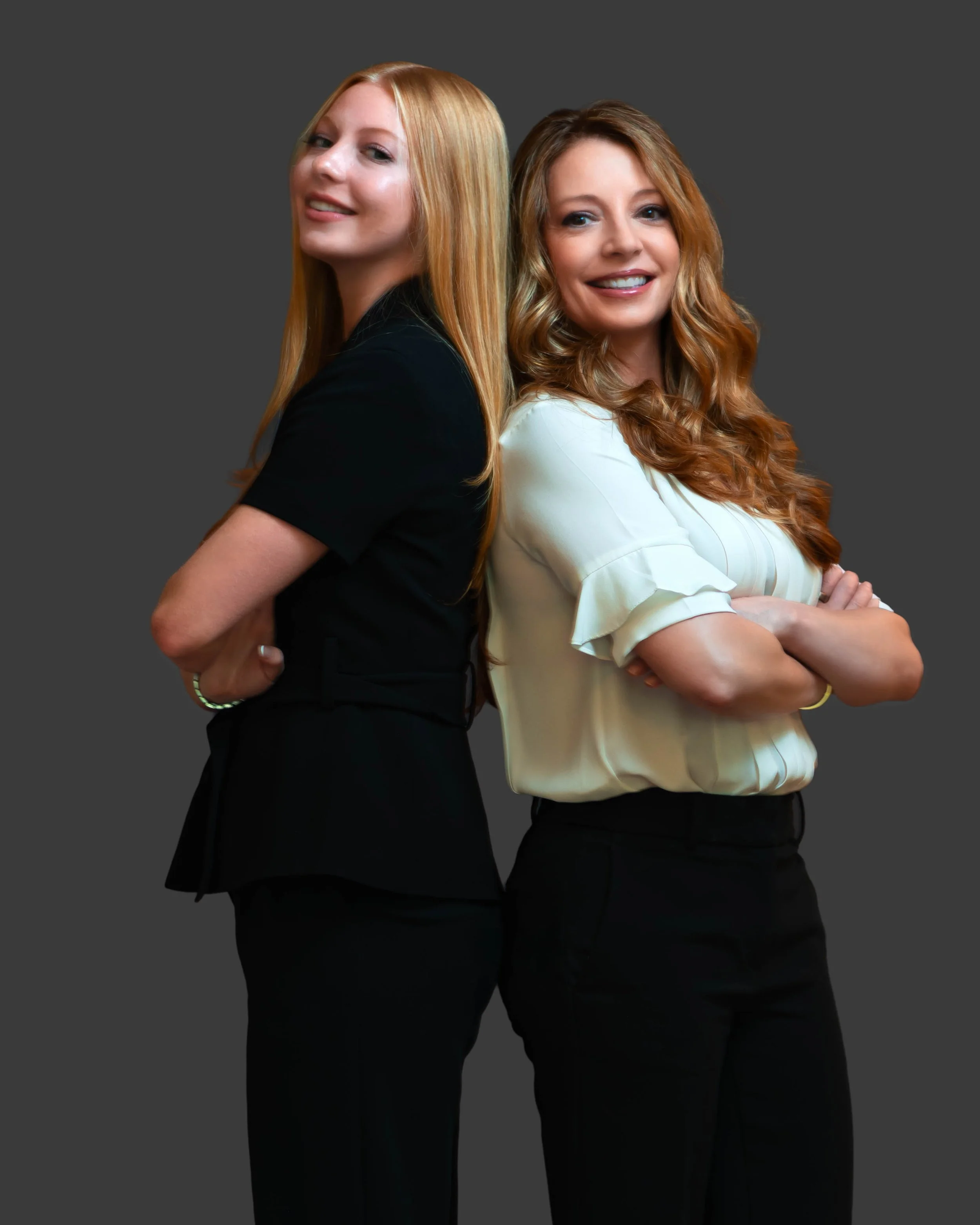 Two women standing back-to-back with arms crossed, smiling, against a dark gray background.