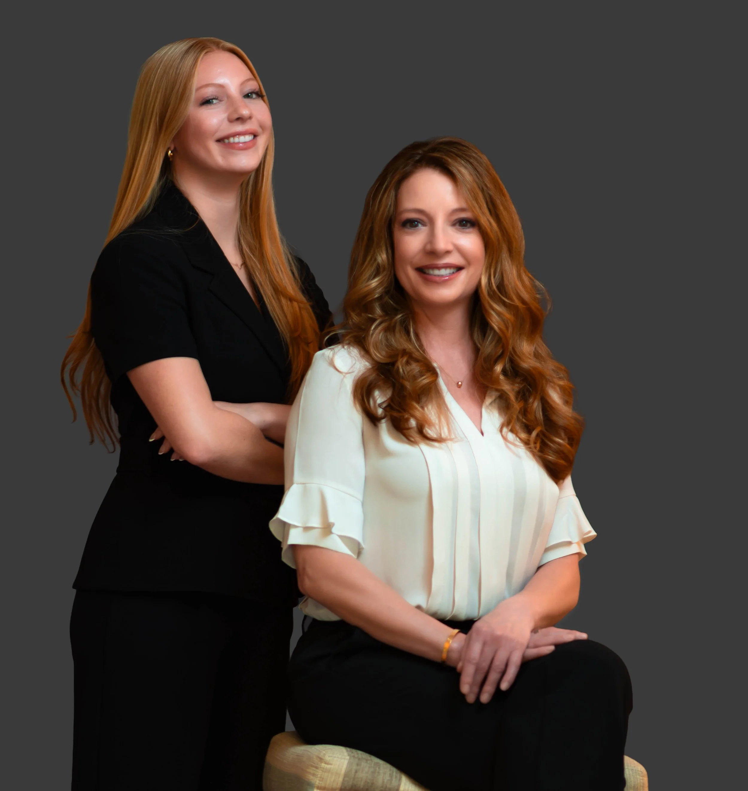 Two women with long red hair posing together against a gray background, one standing behind the seated woman, both smiling.