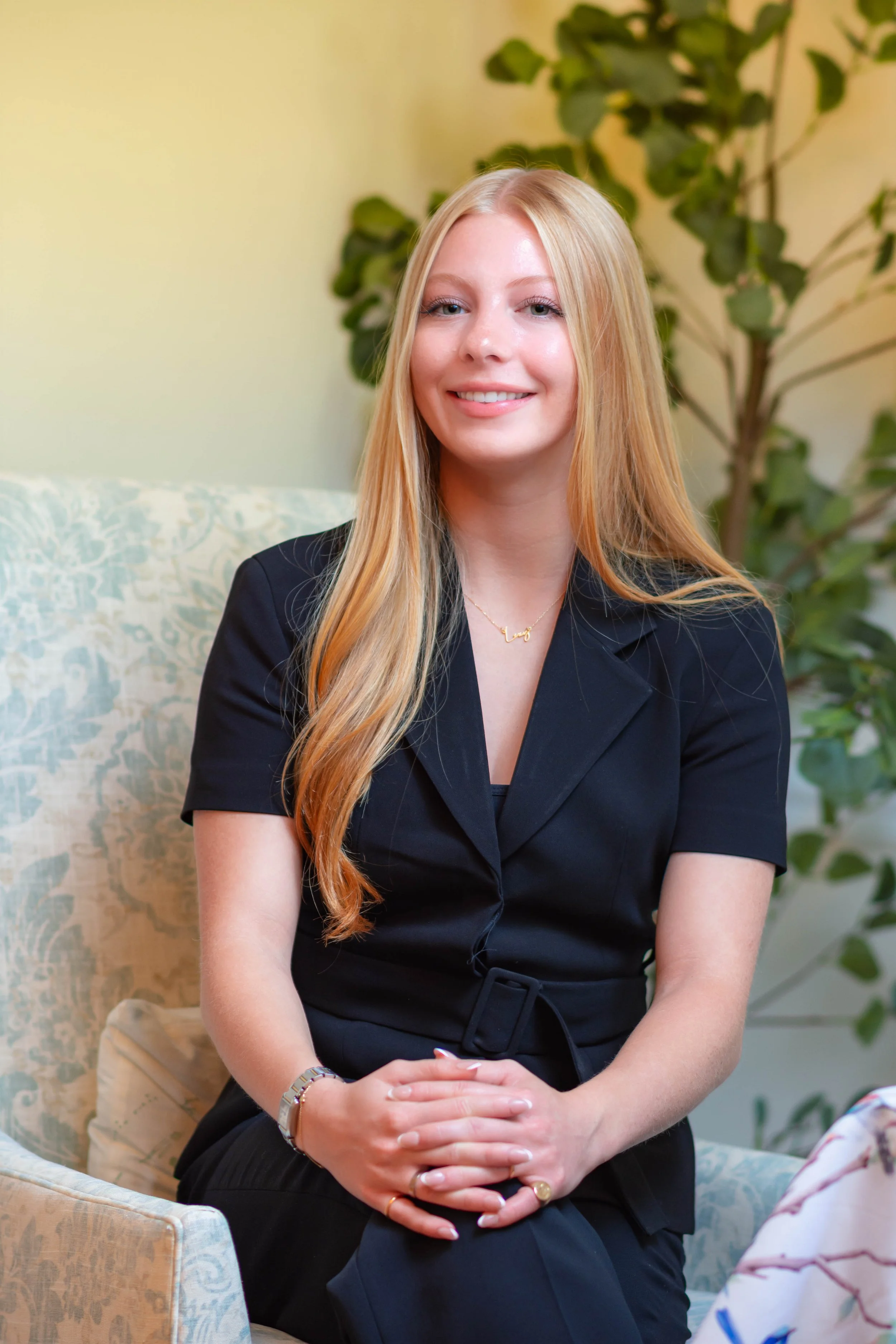 A young woman with long blonde hair, wearing a black dress and a gold necklace, sits on a patterned light-colored chair with her hands clasped in her lap. She is smiling and appears indoors with a green plant in the background.