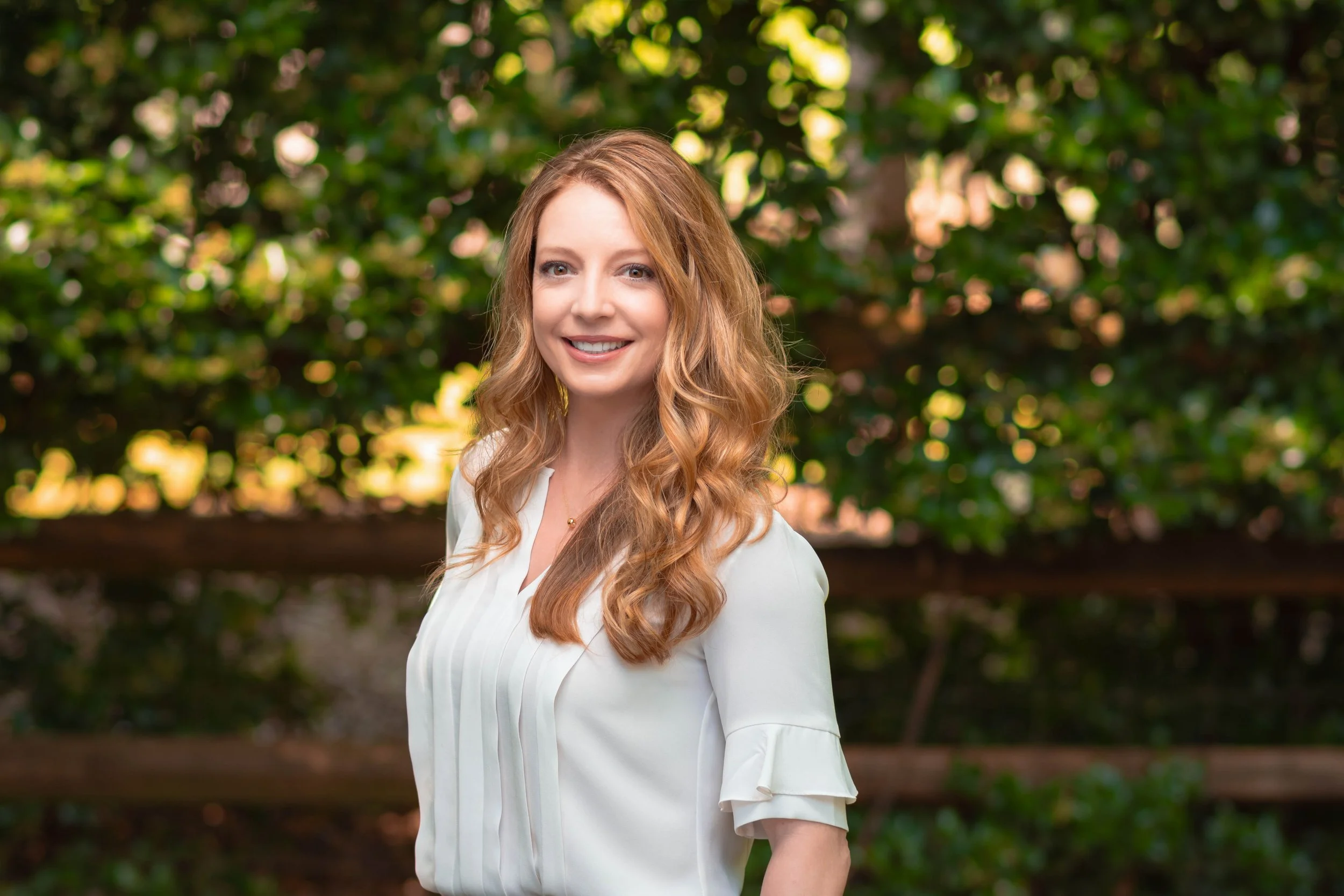 Portrait of a smiling woman with long wavy red hair, wearing a white blouse, standing outdoors with blurred green foliage in the background.