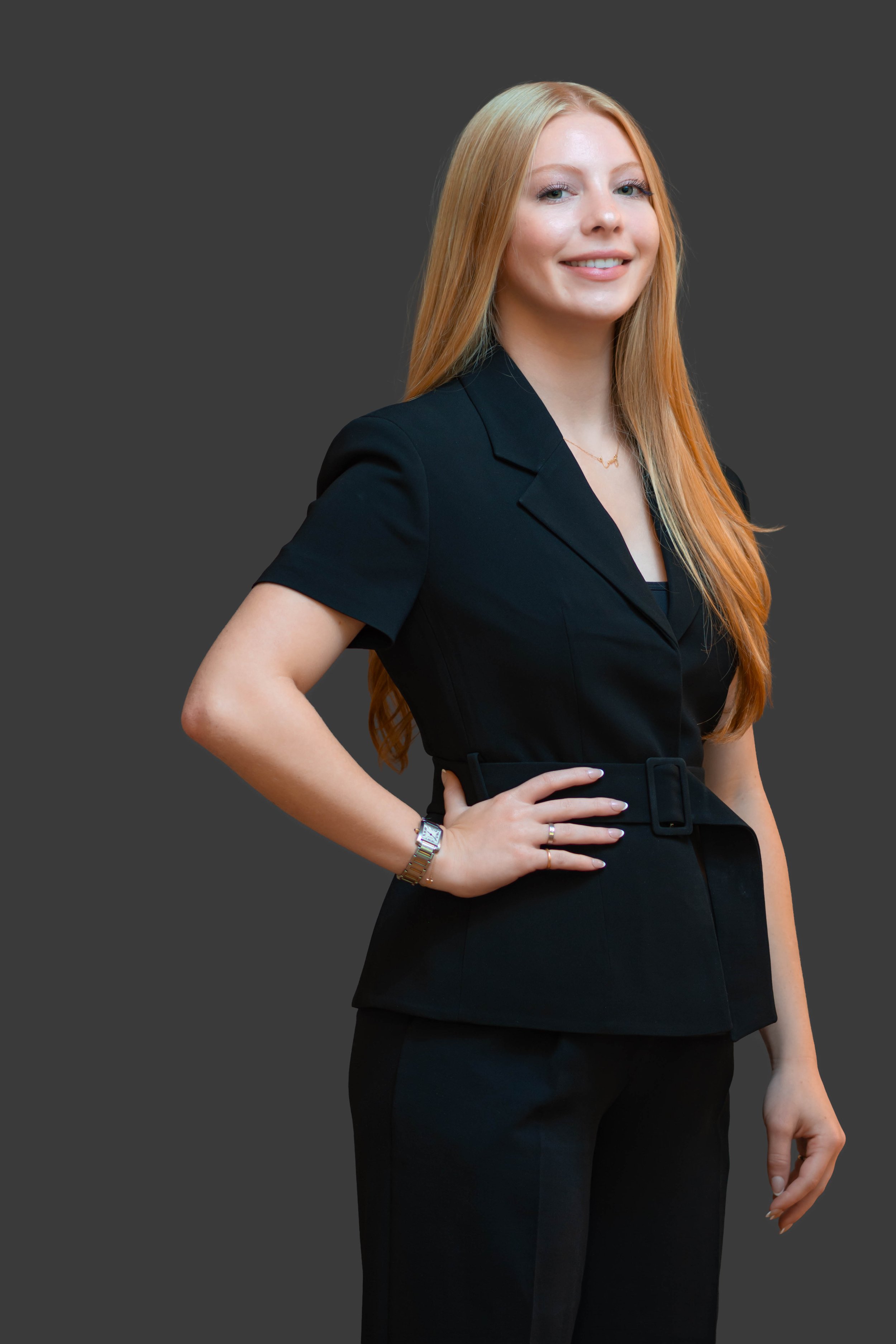 A woman with long red hair, wearing a black blazer and black pants, standing with one hand on her hip and smiling at the camera against a gray background.