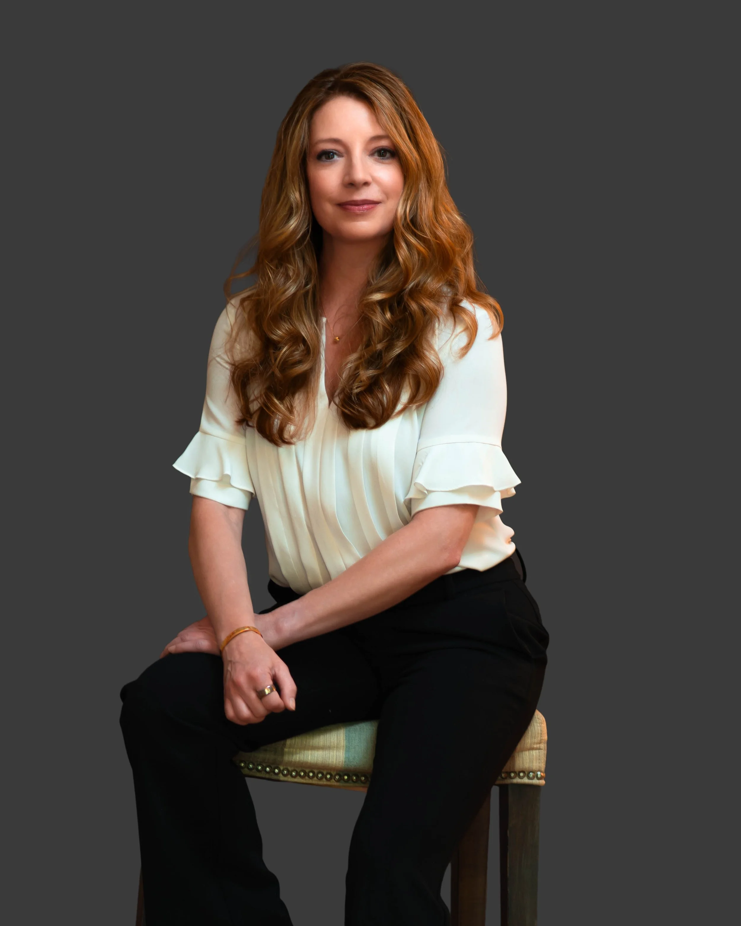 A red-haired woman wearing a cream blouse and black pants sitting on a wooden chair with a dark gray background.