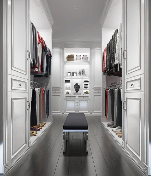 Spacious walk-in closet with white cabinets and shelves, clothing racks with hanging clothes on both sides, a black cushioned bench in the center, and decorative items on the shelves at the far end.