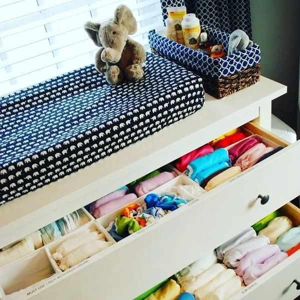 Changing pad with a teddy bear, a basket with baby supplies, and a drawer filled with folded colorful baby clothes.