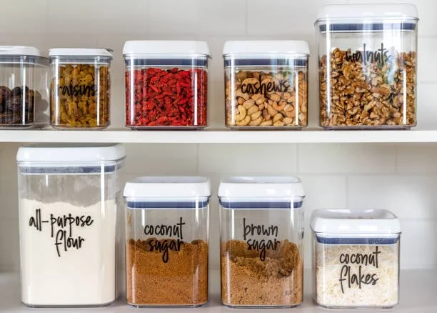Storage jars with labeled dry ingredients on a white kitchen shelf, including raisins, cashews, walnuts, dried apricots, all-purpose flour, coconut sugar, brown sugar, and coconut flakes.