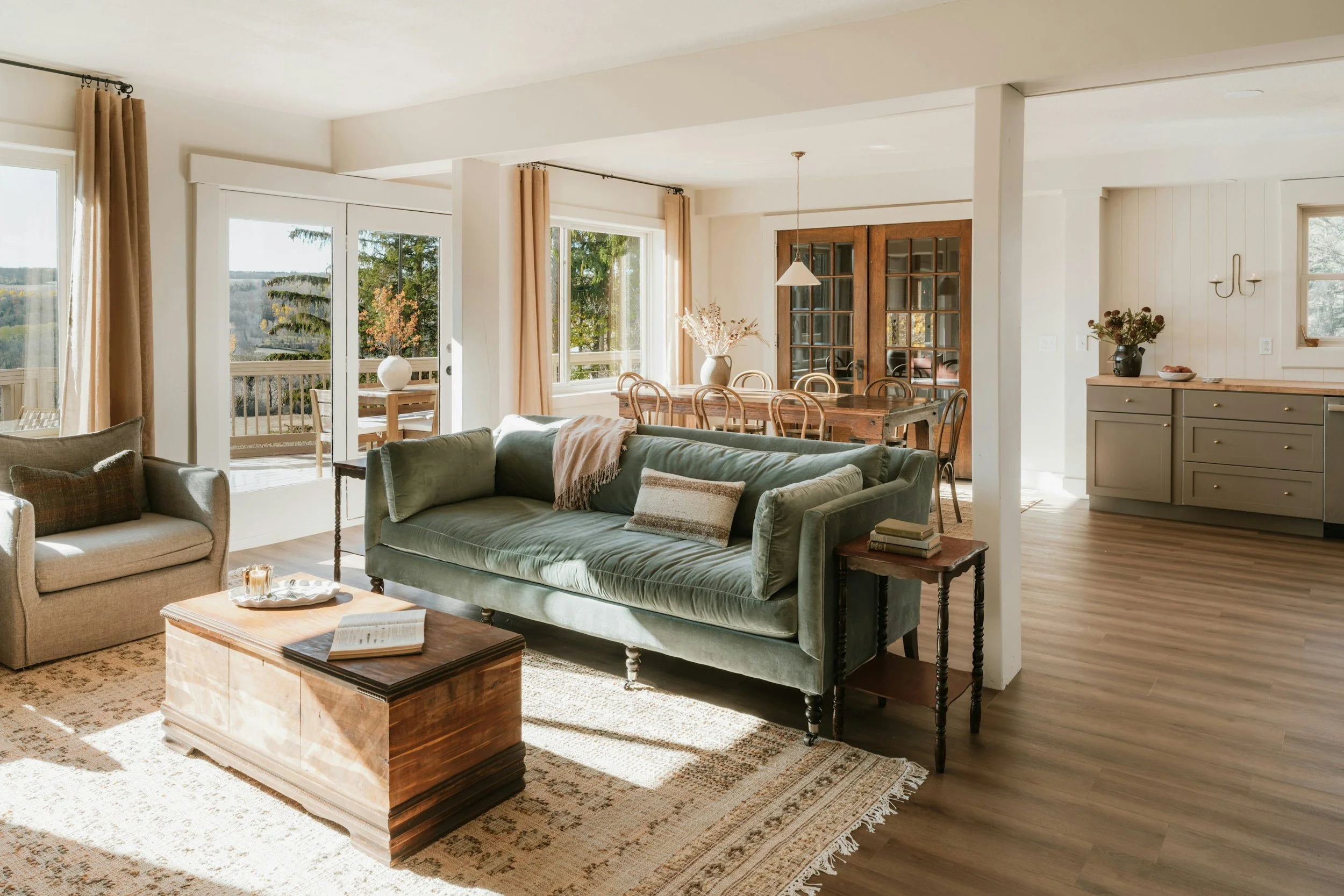 Bright living room with large windows, beige curtains, green velvet sofa, wooden coffee table, and dining area with a wooden table, chairs, and sliding glass door leading to a balcony with an outdoor view of mountains and trees.