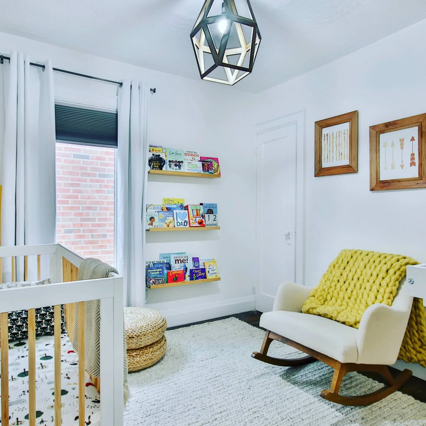 A cozy nursery with white walls, a wooden rocking chair with a yellow knit blanket, a white rug, and floating shelves filled with children's books. There are framed arrow art prints on the wall, a window with white curtains, and a ceiling light fixture.