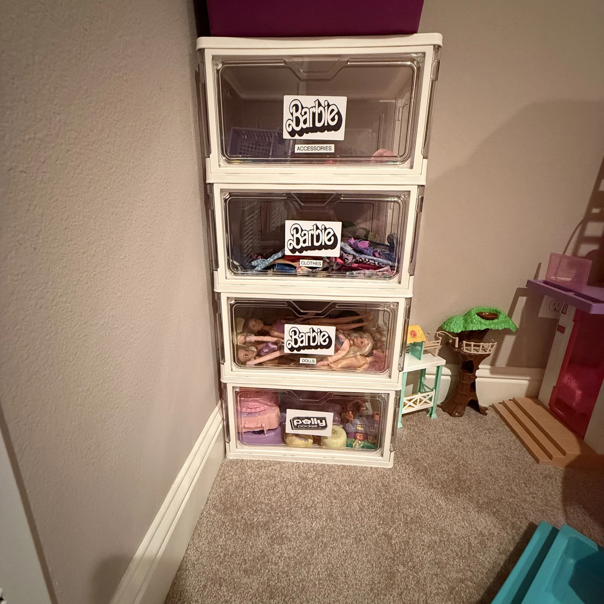 A white storage unit with five transparent plastic drawers labeled 'Barbie Accessories', 'Barbie Dolls', and 'Puffy Pocket'. The drawers contain various Barbie toys and accessories. To the right, there is a small white and green table with a dollhouse or playset on top, and a lavender chair nearby.