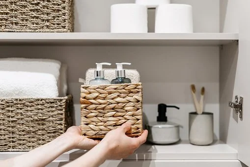 A hand placing a woven basket holding two soap dispensers into a white cabinet with bathroom items, including towels and brushes.