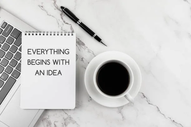A flat lay of a workspace with a laptop, a notepad with the quote 'Everything begins with an idea,' a black pen, and a cup of black coffee on a white marble surface.
