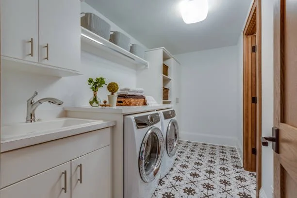 Laundry room with white cabinets, a sink, and a washing machine and dryer. There are decorative items on top of the appliances and open shelving with baskets. The floor has patterned tiles and a wooden door is partially open.