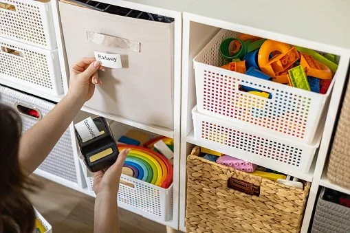 Child sorting toy blocks and rainbow-colored toys in a white storage unit with labeled and open baskets.