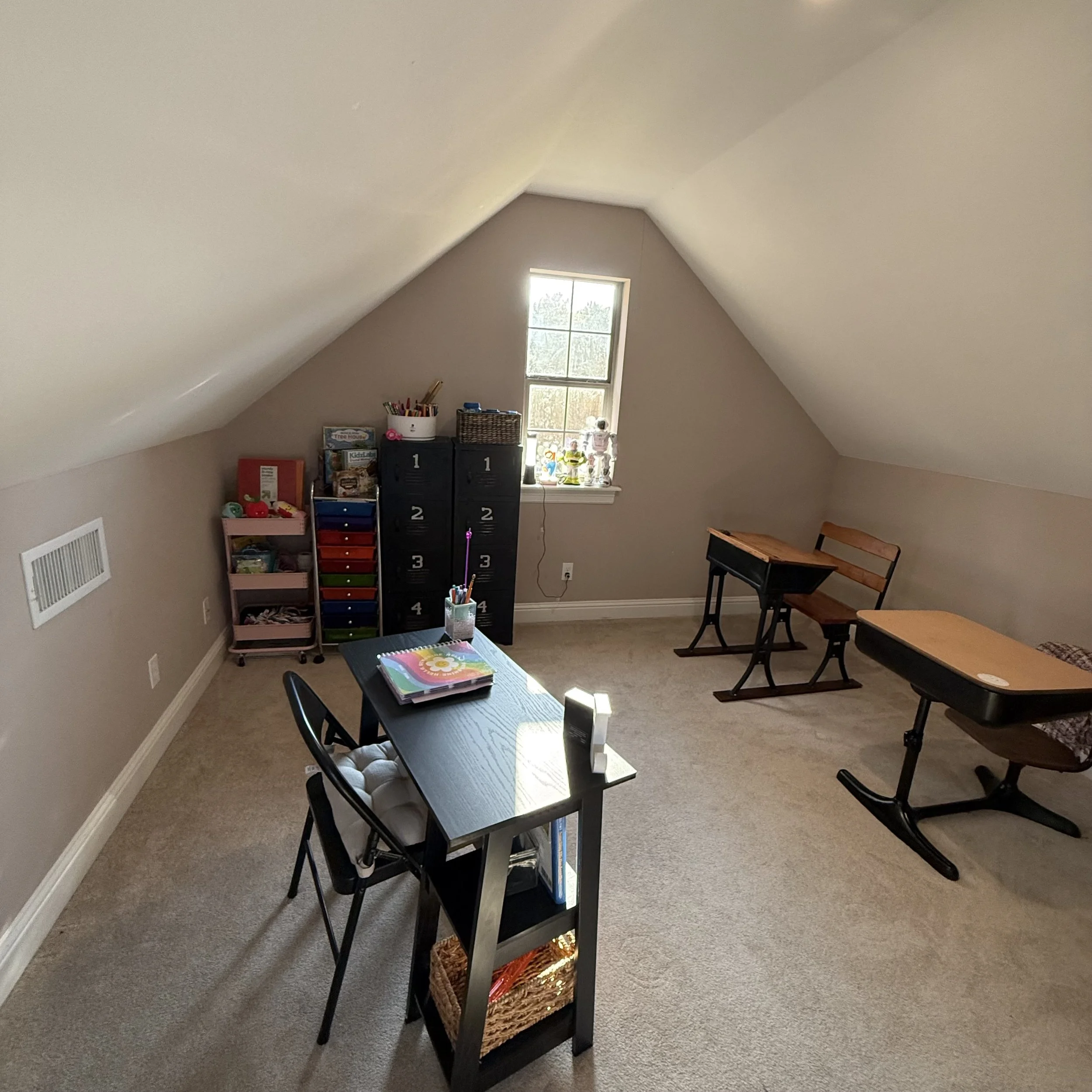A room with sloped ceilings and a window at the back wall, furnished with a small table, chairs, and storage units filled with toys and books.