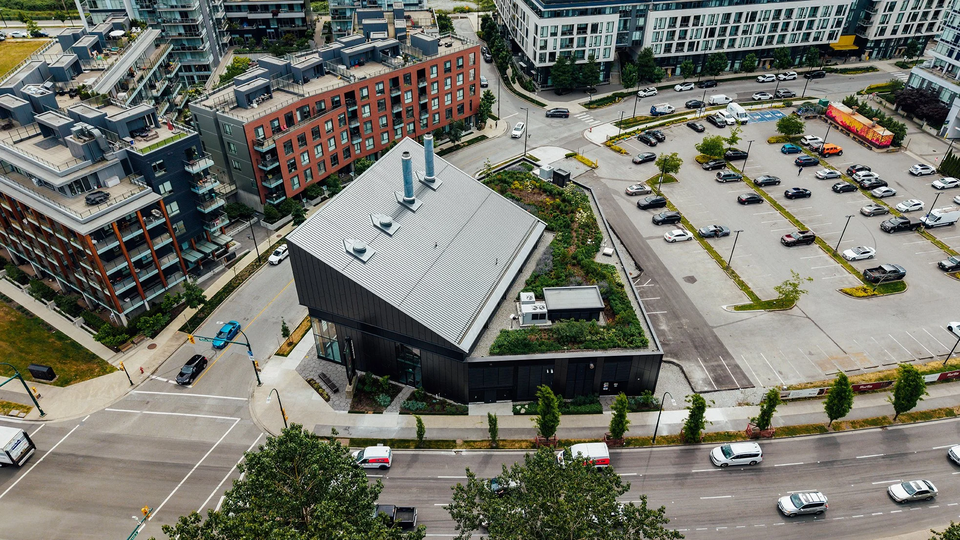 Aerial view of a city block with modern apartment buildings, a parking lot with cars, and a building with rooftop garden and chimneys.