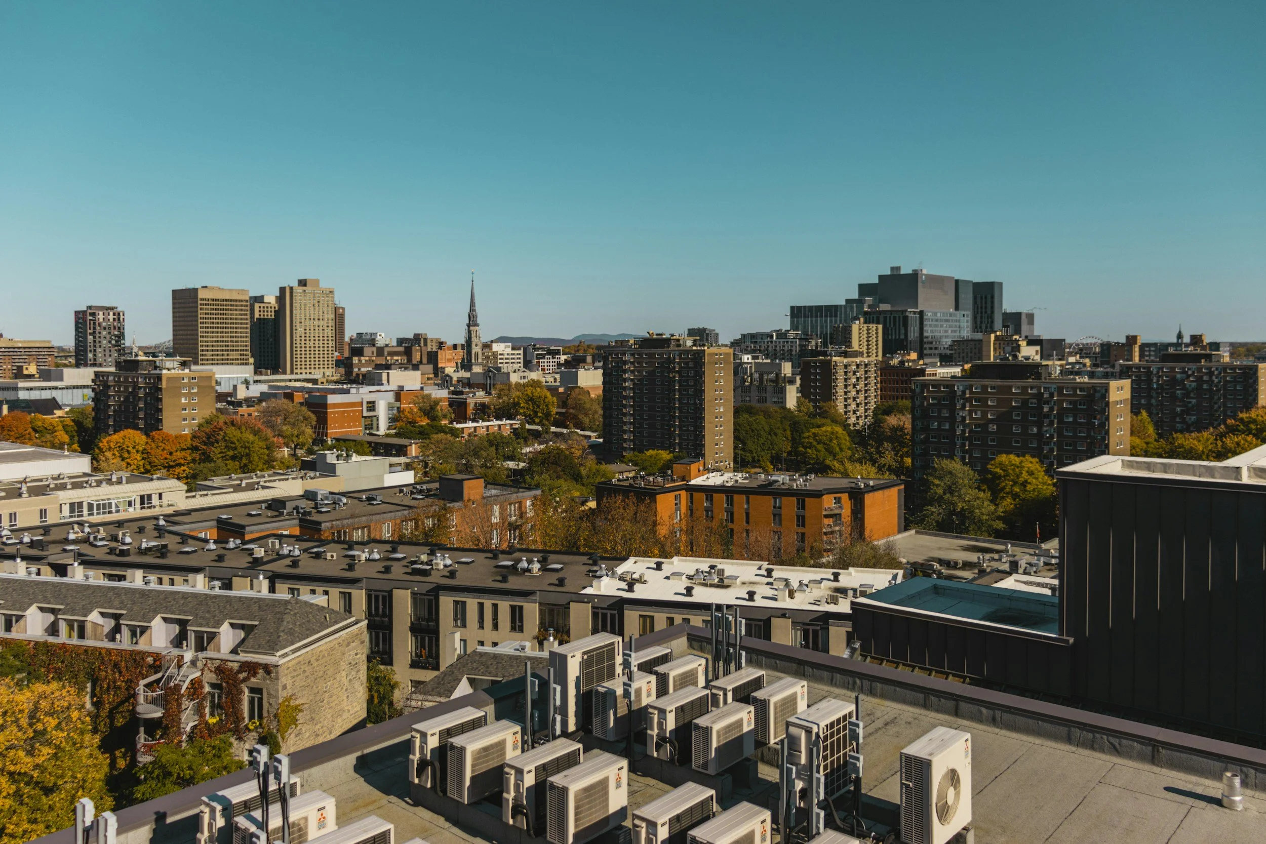 Vue d'une ville avec des immeubles modernes, quelques arbres aux couleurs d'automne, un ciel dégagé et une rangée de climatiseurs ou de systèmes de ventilation sur le toit d'un bâtiment.