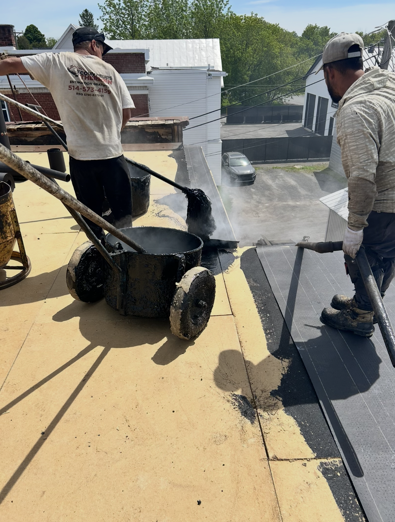 Deux ouvriers travaillent sur la toiture d'un bâtiment résidentiel, appliquant une couche de membrane d'étanchéité. L'un d'eux utilise une buse pour vaporiser un produit, tandis que l'autre tient une lance pour étendre la membrane. Le chantier est en extérieur, avec des maisons et des arbres en arrière-plan.