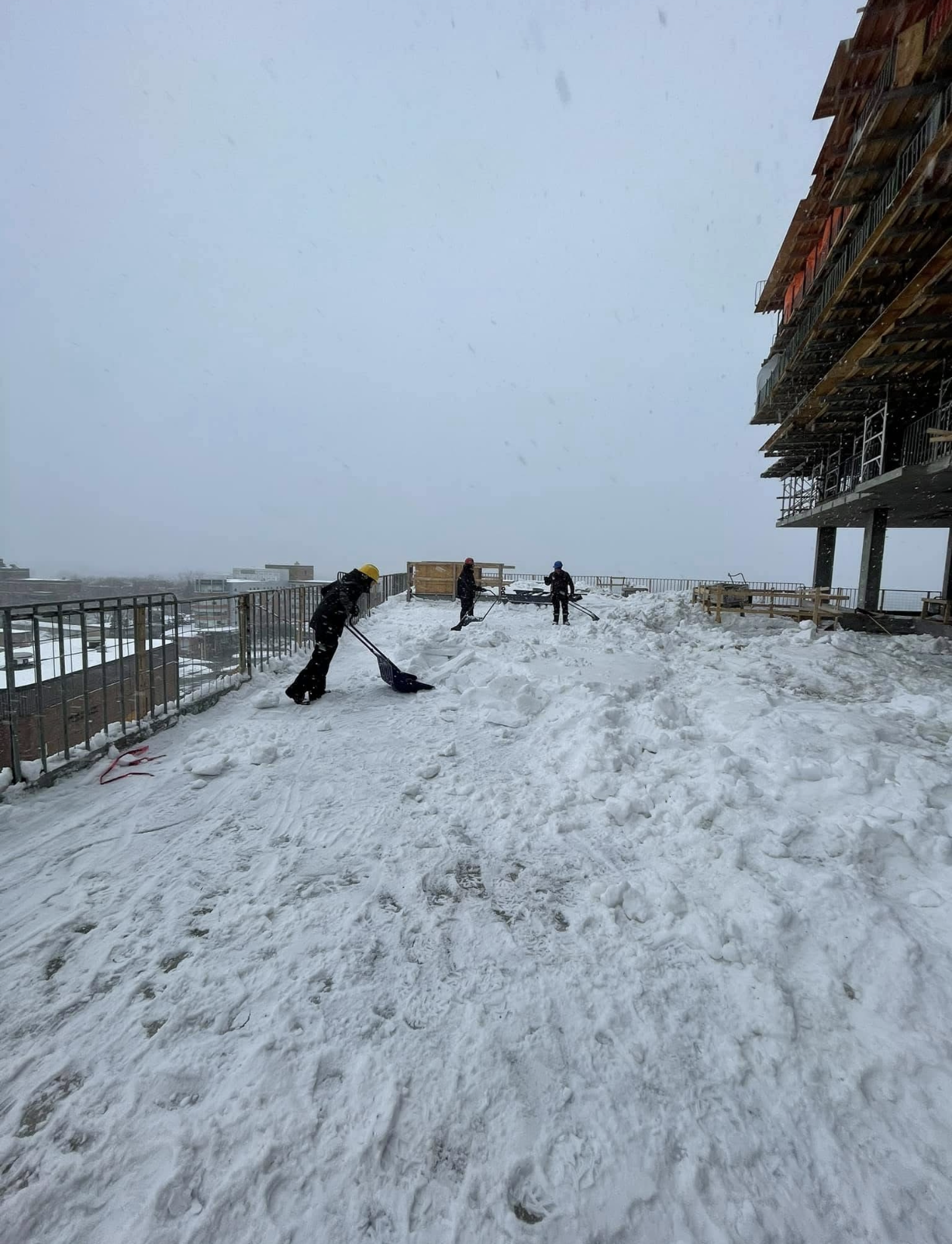 Travailleurs déneigeant un terrain enneigé sur un toit, avec un bâtiment en construction à droite et un ciel gris et brumeux.