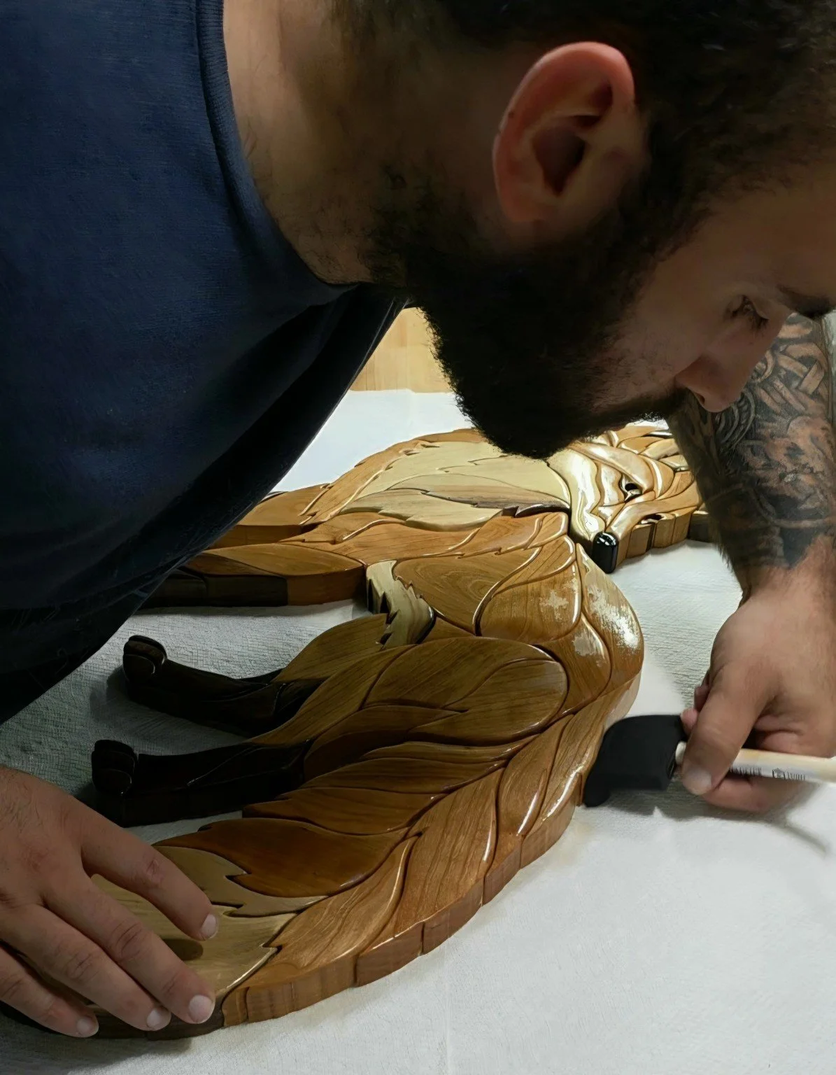A man with a beard and tattoos is oiling a wooden fox sculpture with a foam brush. The sculpture appears to be an artistic piece made of polished wood, resting on a white surface.