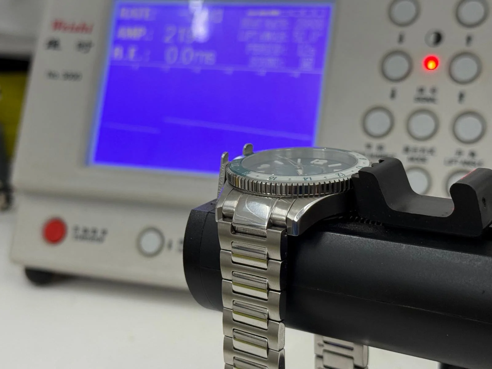 Un orologio con cinturino in metallo appoggiato su un supporto nero, con apparecchiature elettroniche e uno schermo blu sullo sfondo.