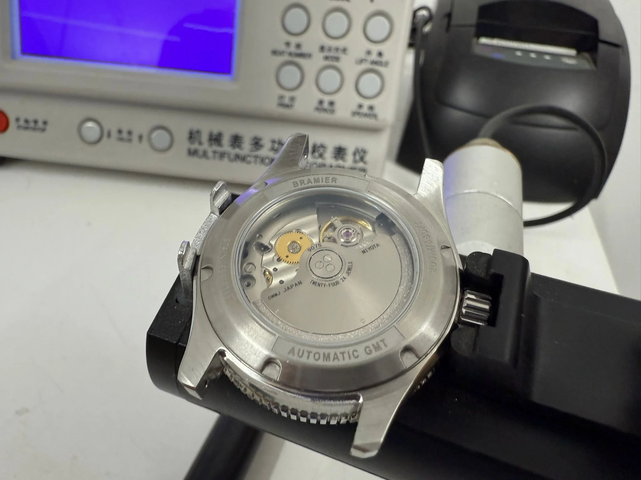 Un orologio meccanico con movimento automatico, rimosso dal suo cinturino, posizionato su una superficie bianca, con un apparecchio di misurazione elettronico e una lampada UV sullo sfondo.