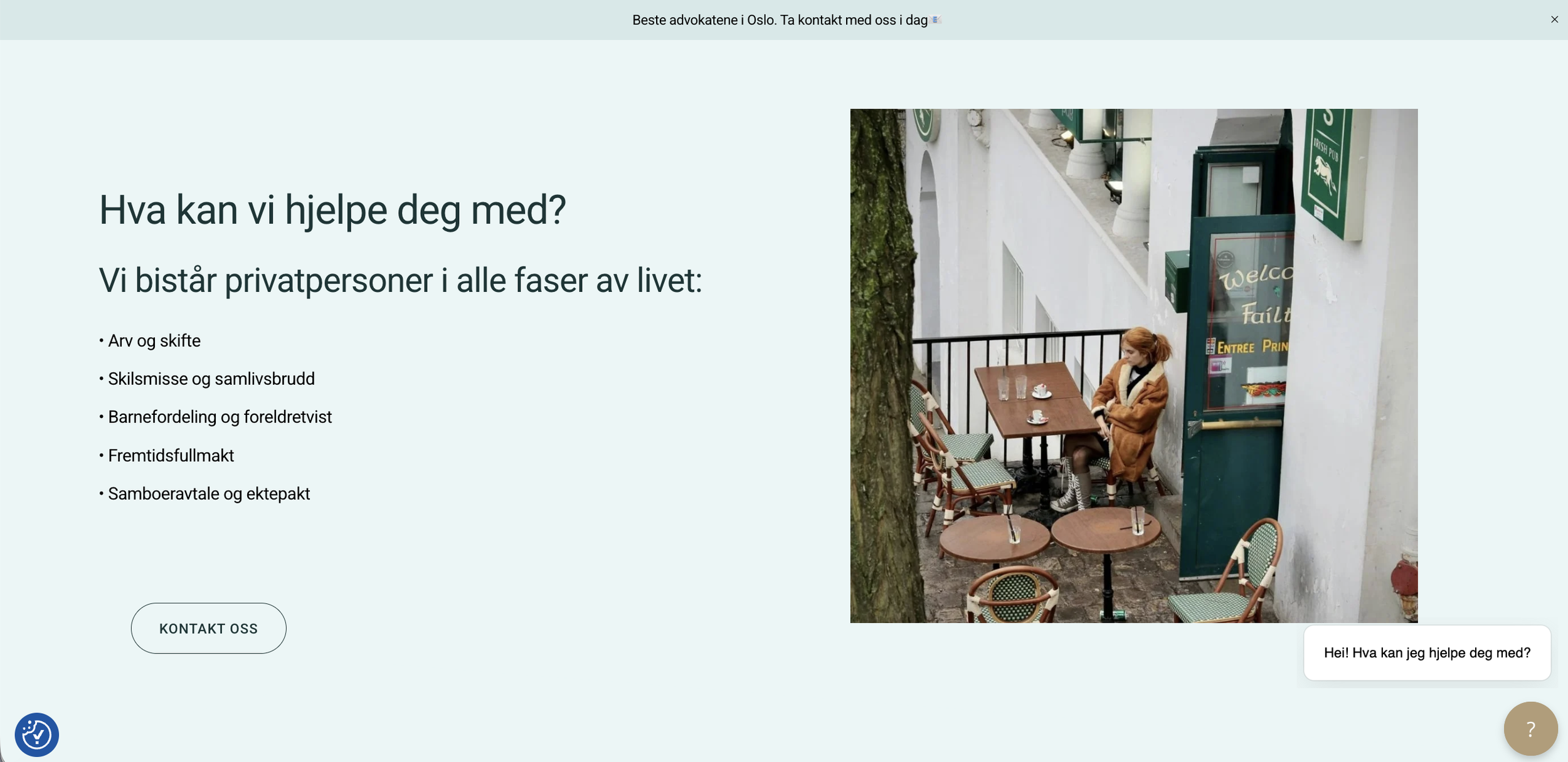 En person med rødt hår som sitter alene ved et bord utendørs på en restaurant eller kafé, omgitt av stoler og bord, ved siden av en hvit bygning og et tre.