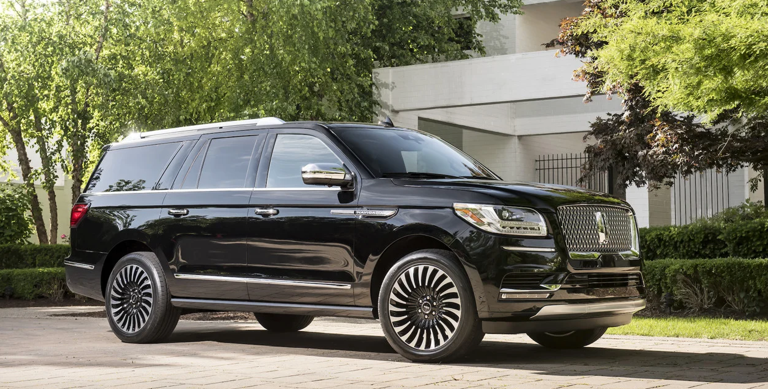 A black Lincoln Navigator SUV parked on a suburban street with green trees and a modern white building in the background.