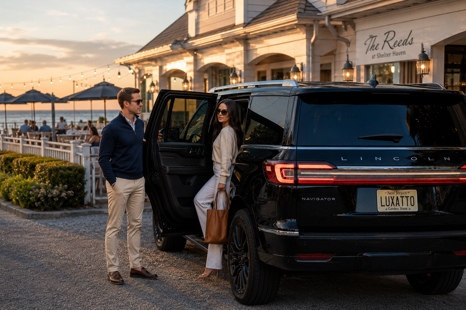 A man and woman near a black Lincoln Navigator SUV outside a restaurant by the beach at sunset. The woman is stepping out of the vehicle, carrying a brown bag, and both are wearing sunglasses.