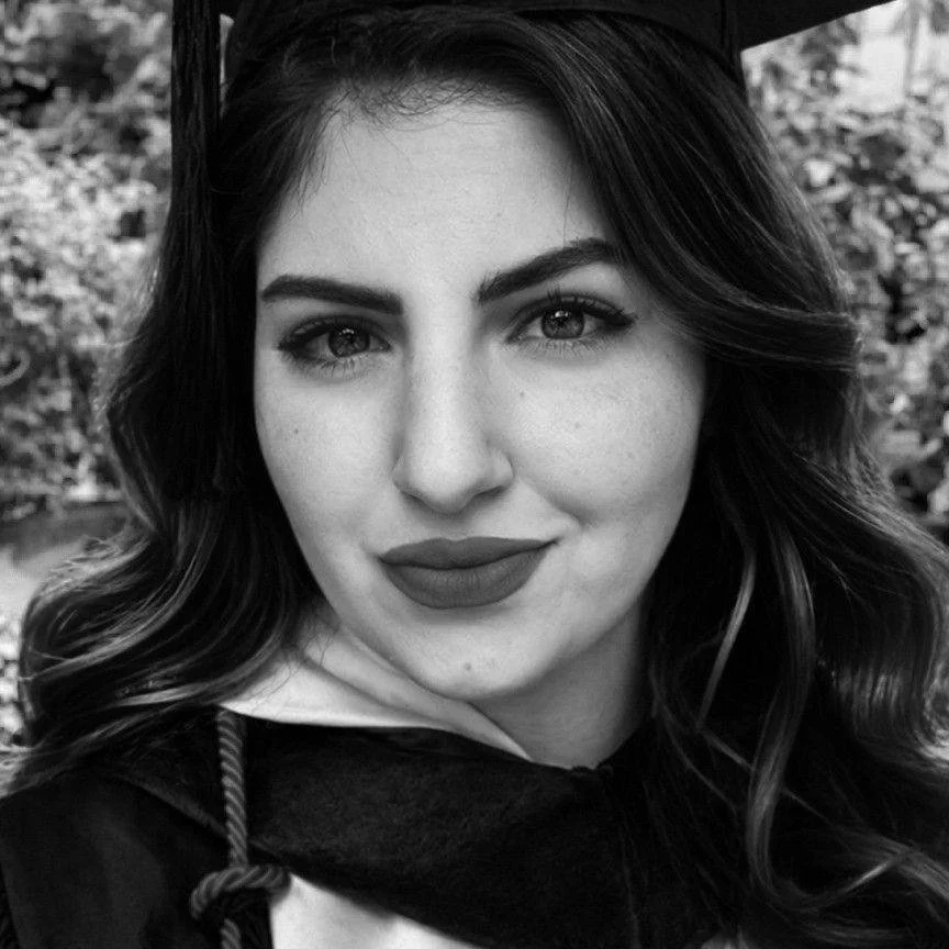 Close-up of a young woman with wavy hair, wearing a graduation cap and gown, outdoors background.