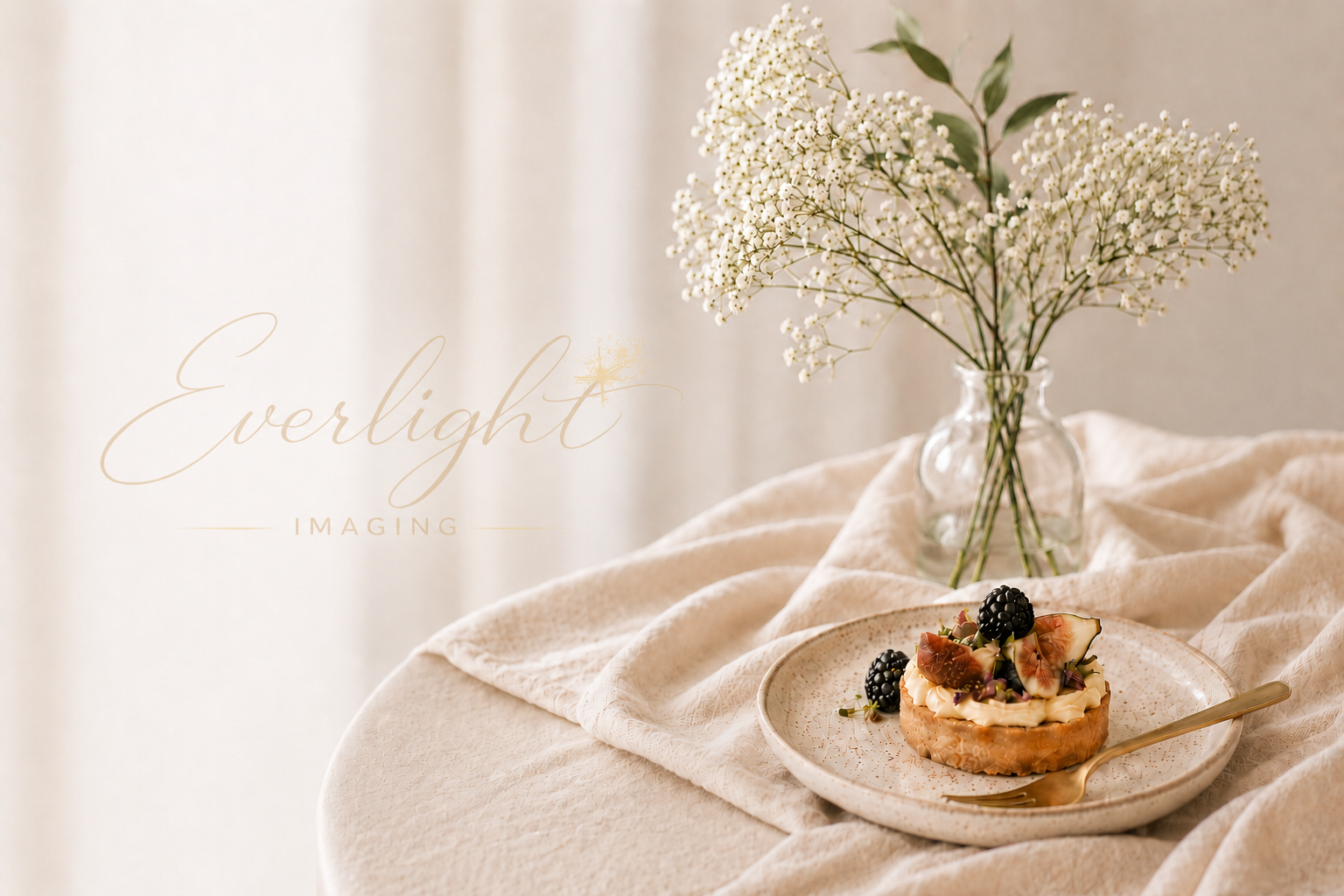 A slice of pastry topped with blackberries and figs on a speckled plate with a gold fork beside it, and a vase with white baby's breath flowers on a beige tablecloth.