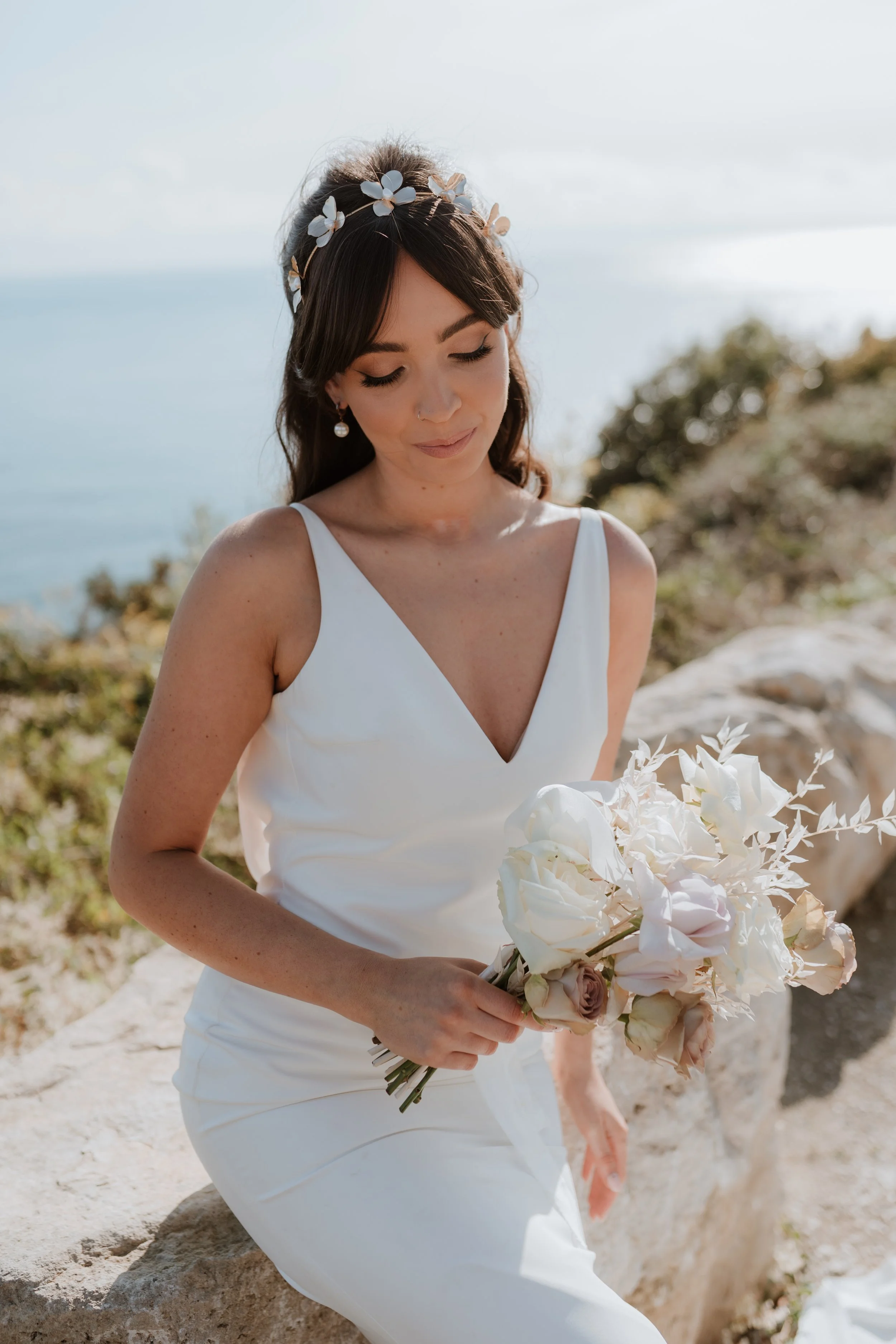 A woman dressed in a white dress, holding a bouquet of white and blush flowers, standing outdoors with a blurred natural background.
