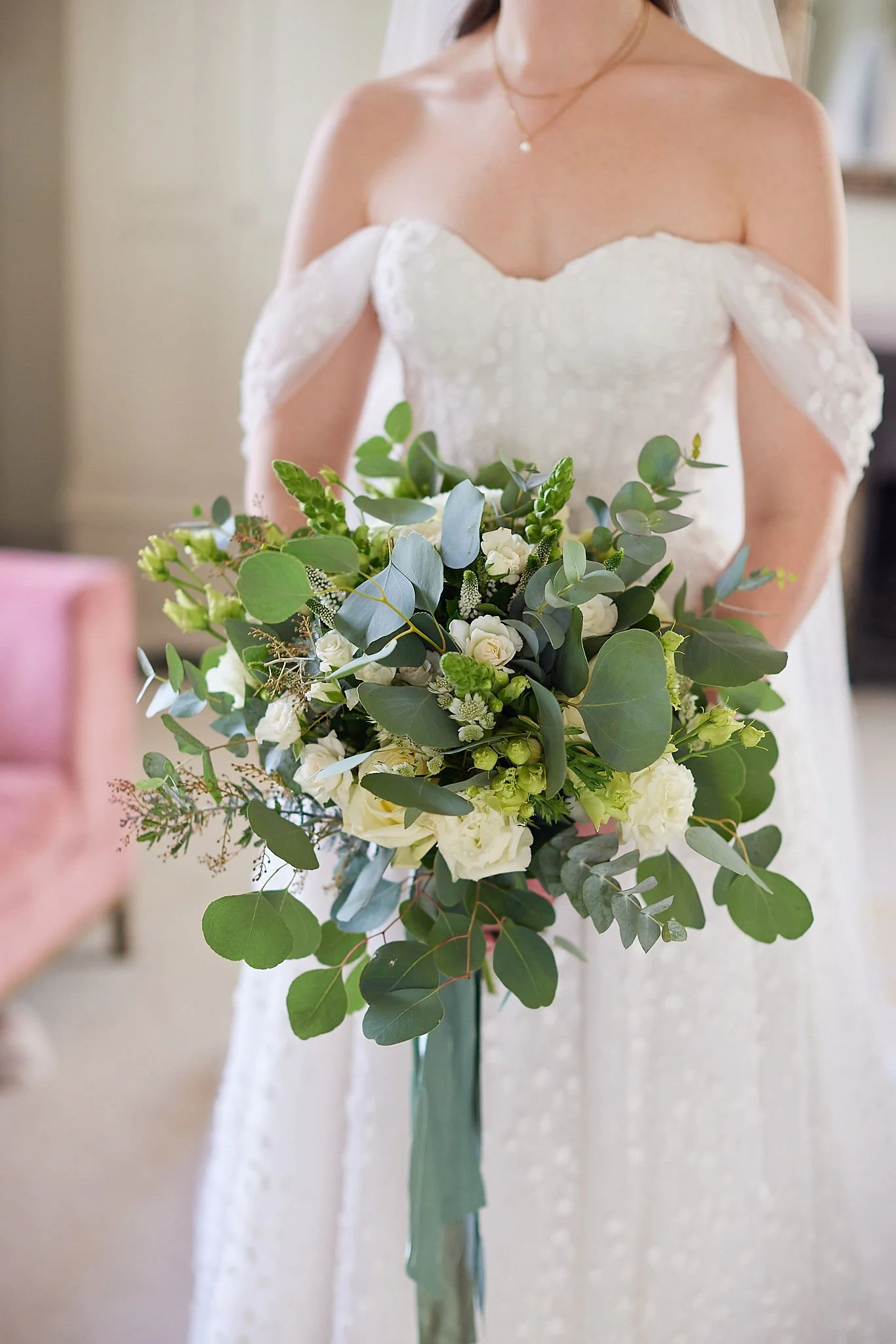 A woman in a white off-the-shoulder wedding dress holding a bouquet of white flowers and green foliage.