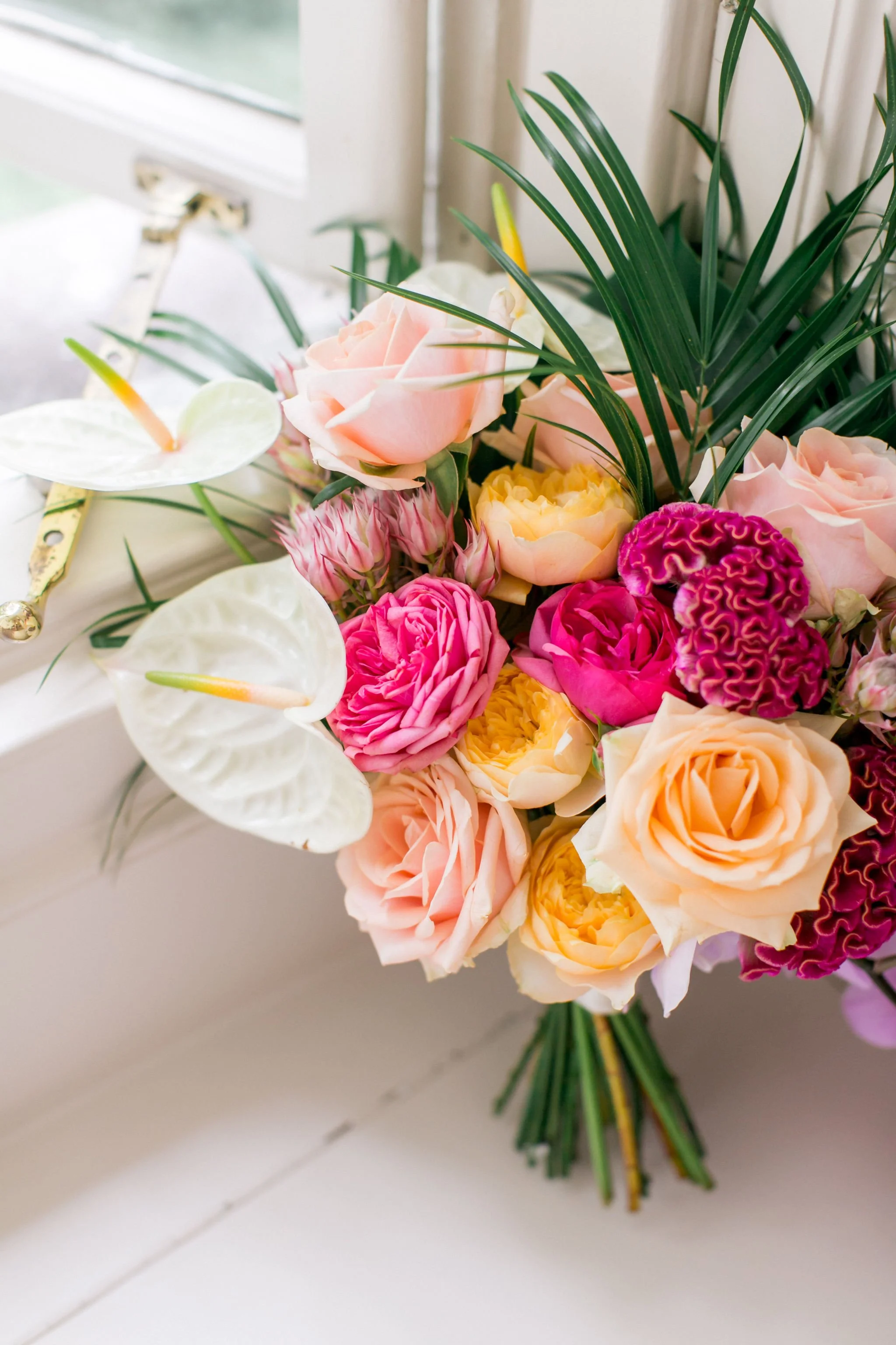 A bouquet of pastel-colored flowers, including pink roses, white calla lilies, and purple and yellow carnations, with green foliage, arranged near a window with white curtains.