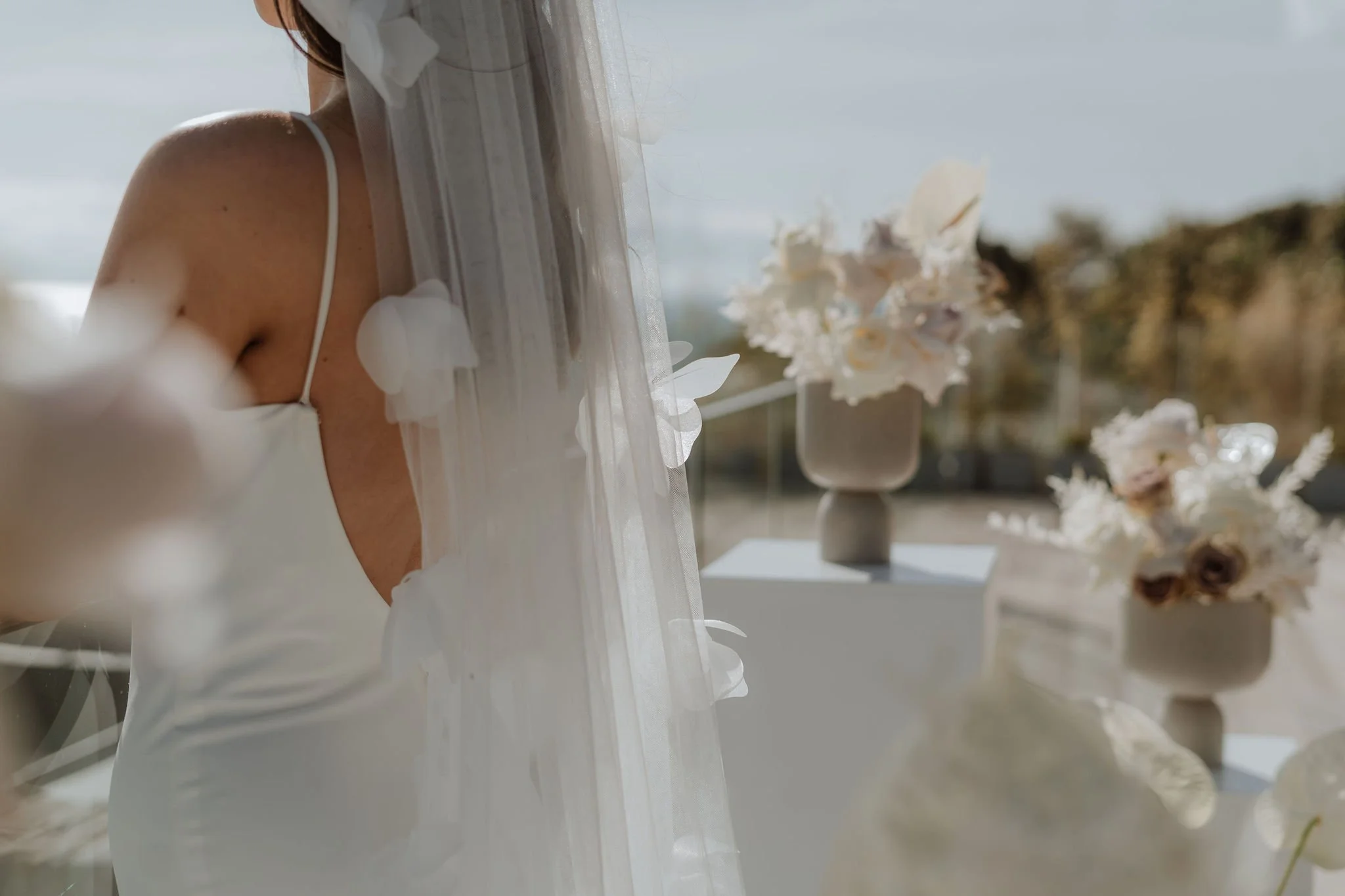 A woman wearing a white dress with thin straps and a veil, with her face not visible, standing outdoors with blurred white flowers in the background.
