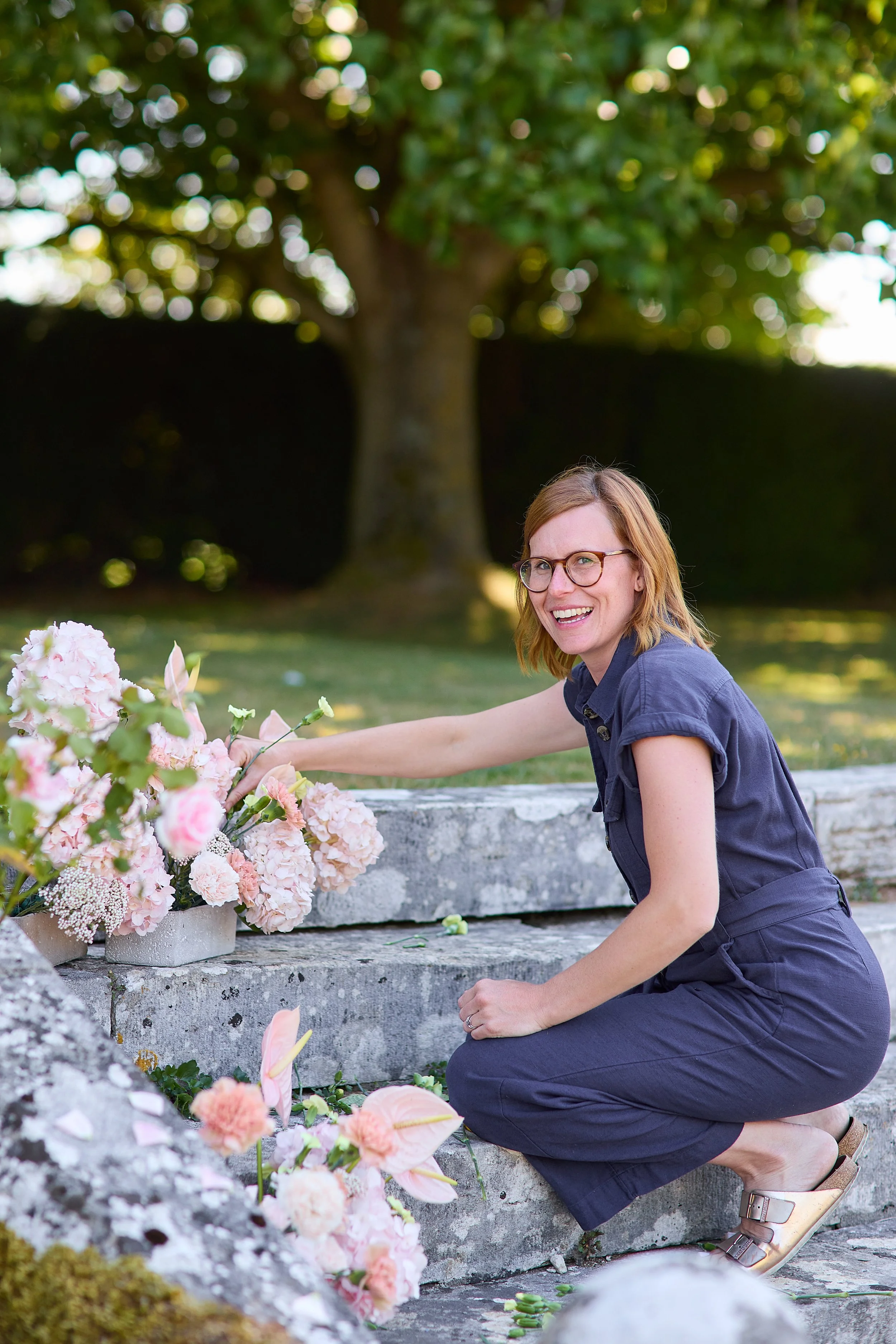 A woman with glasses and shoulder-length hair in a navy dress, kneeling outdoors near a gray stone monument or bench, arranging pink and white flowers at a memorial site with trees in the background.