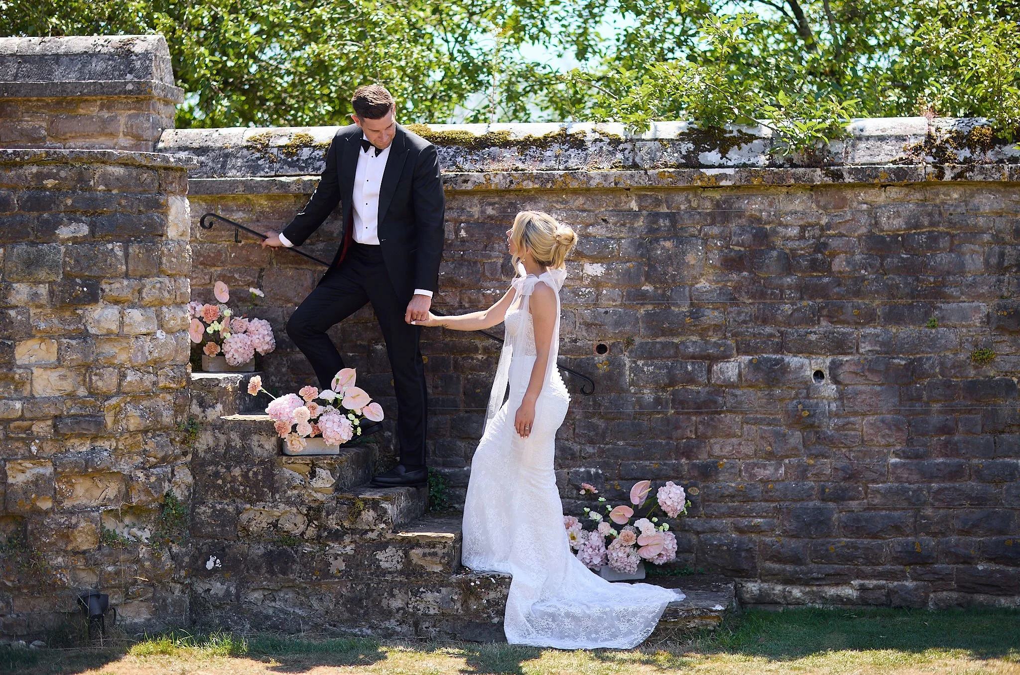 A groom in a black tuxedo holding a cane, kneeling on a stone step, holding the hand of a young girl in a white dress, in front of a weathered brick wall decorated with pink and cream flowers, outdoors under trees.