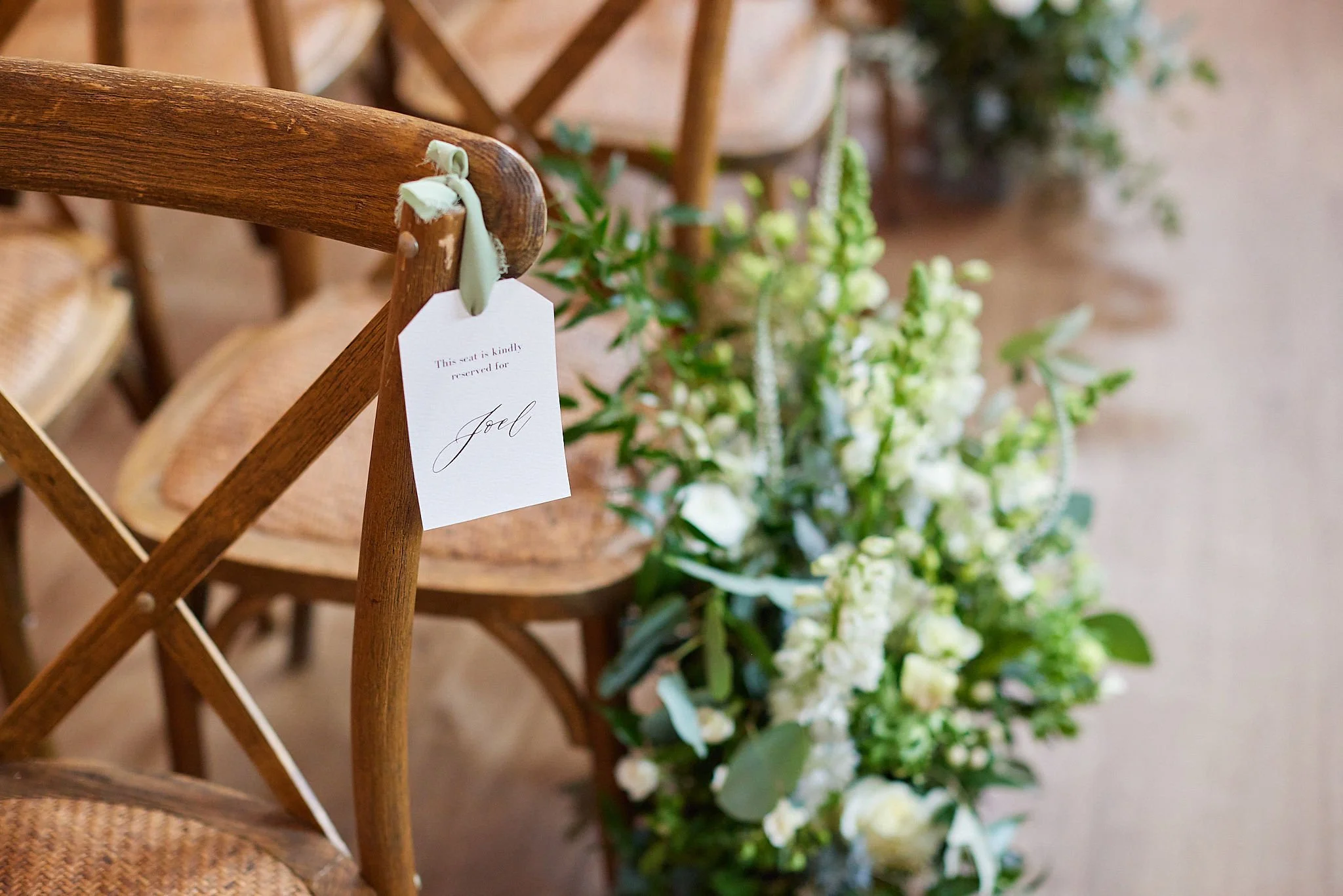 Close-up of wooden chairs arranged in a line with wedding or event decorations, including a white tag that says 'This seat is kindly reserved for Jill', and green floral arrangements in the background.