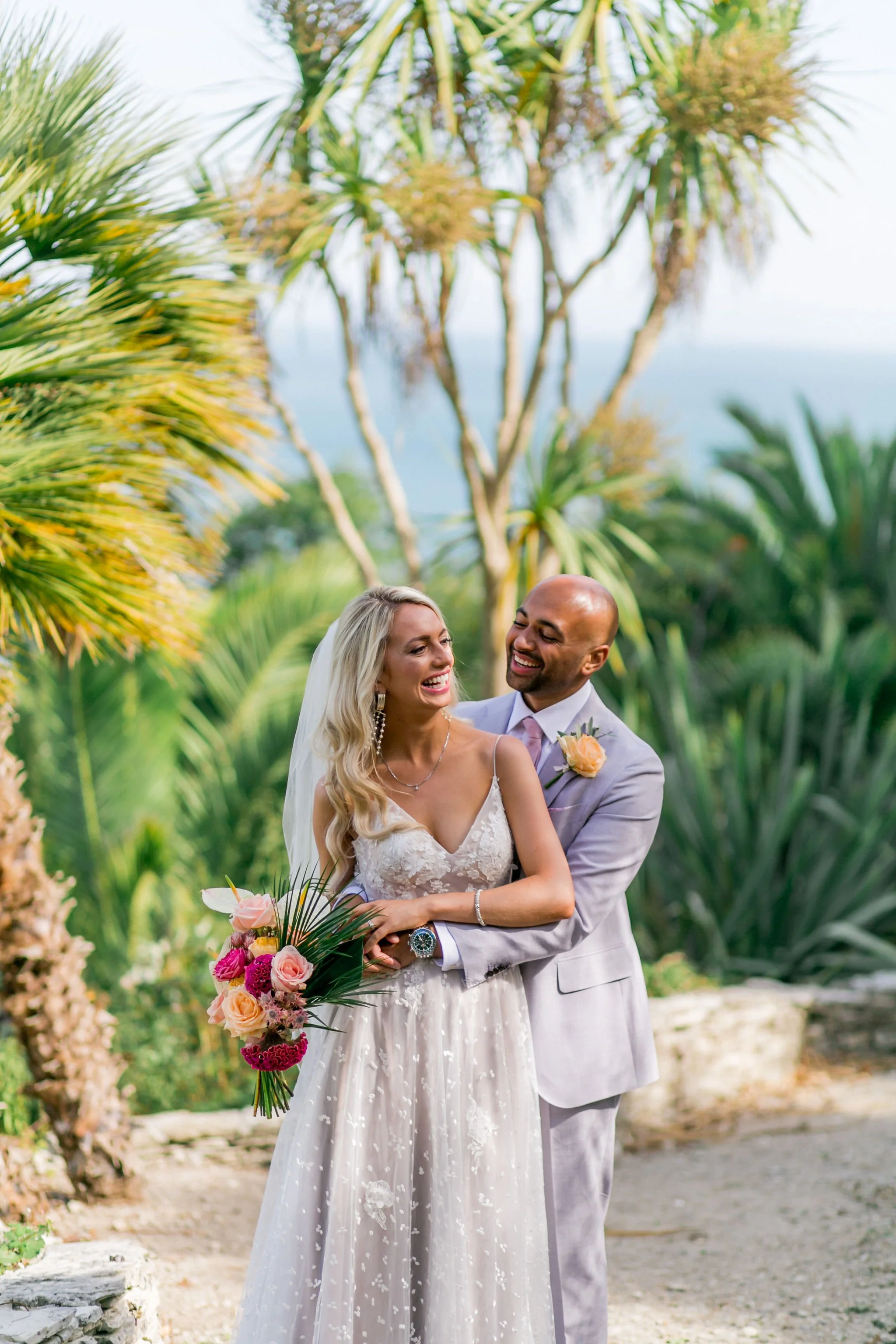 A bride and groom smiling and embracing during their wedding ceremony outdoors surrounded by lush green tropical plants and trees.
