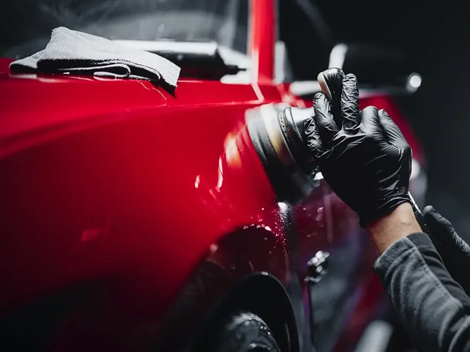 A person wearing black gloves polishing a red car with a power buffer in an auto body shop.